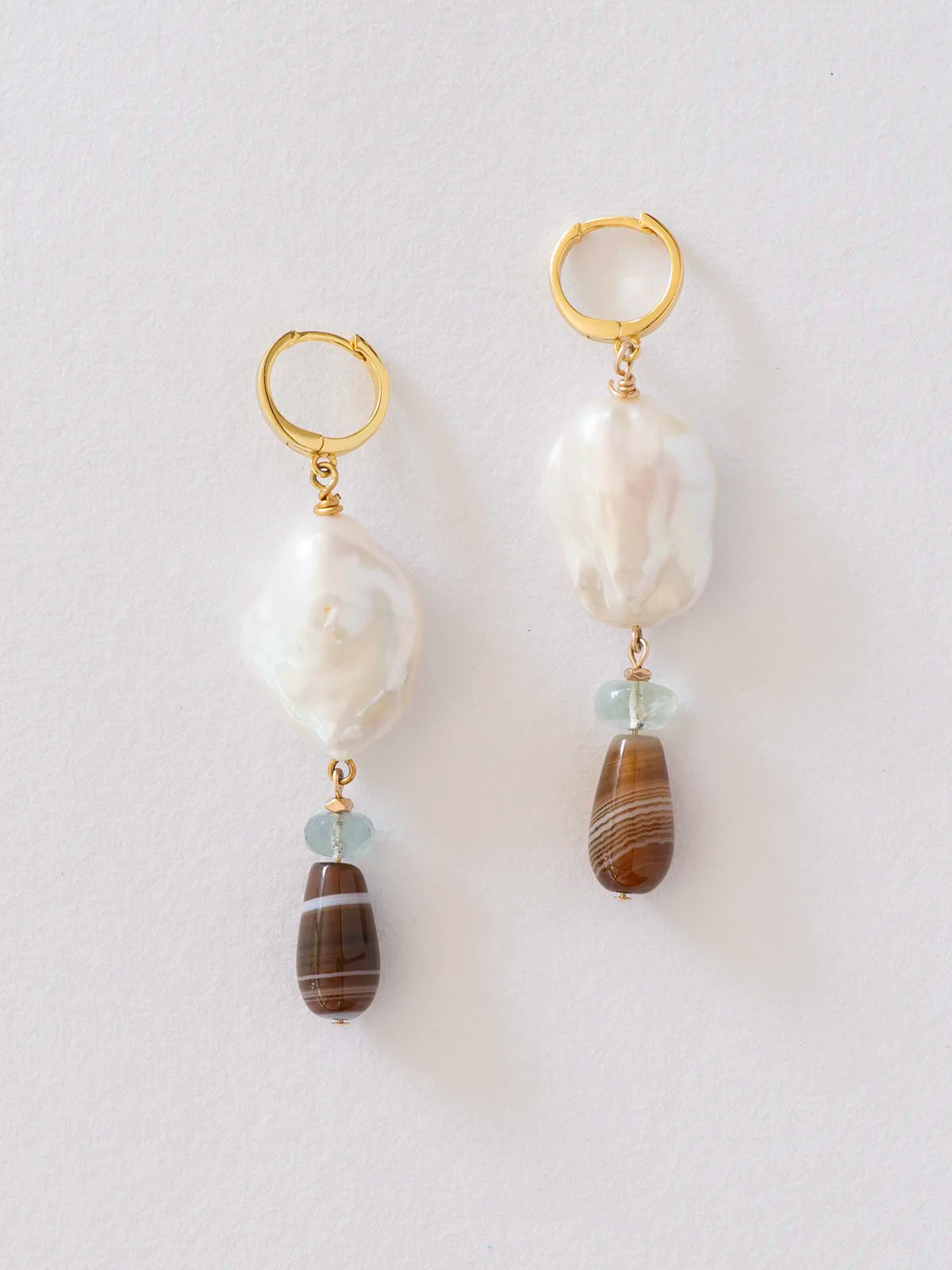 A pair of gold hoop drop earrings featuring a large white baroque pearl, a small light blue bead, and a brown striped agate bead, displayed against a clean white background.