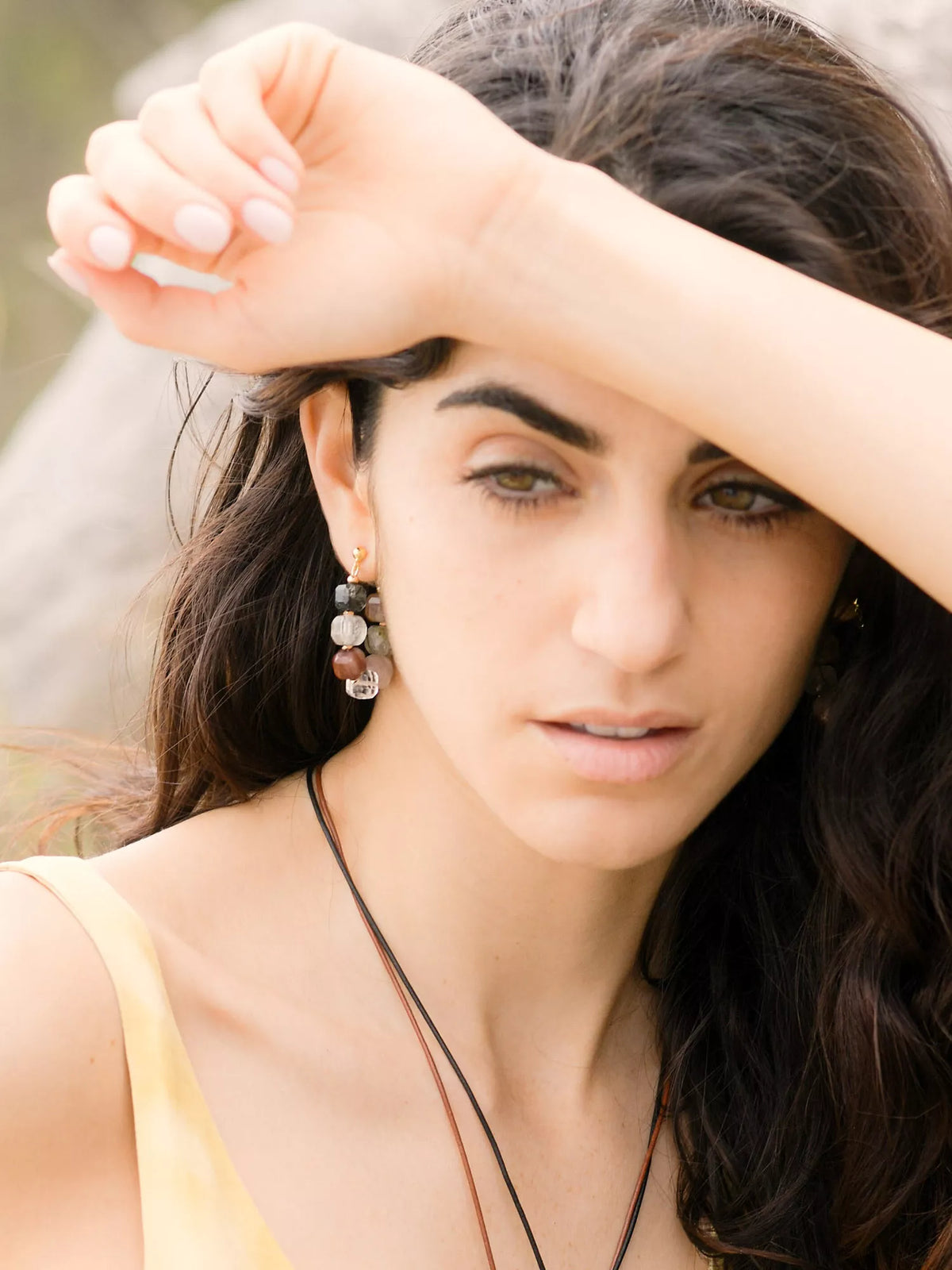 Woman with dark hair and earrings, wearing a yellow top, outdoors.
