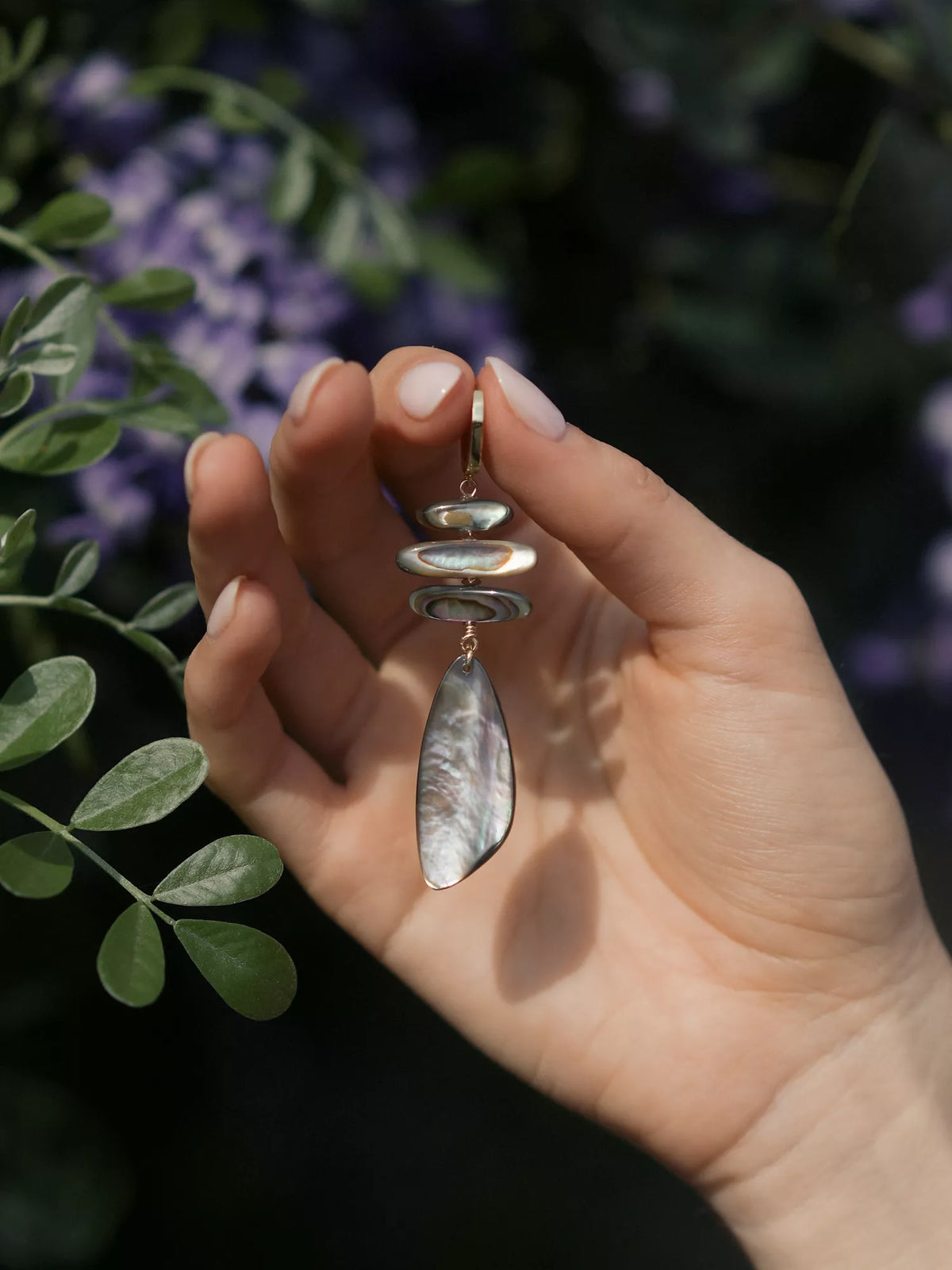 Hand holding an abalone earring with a natural background of leaves and flowers