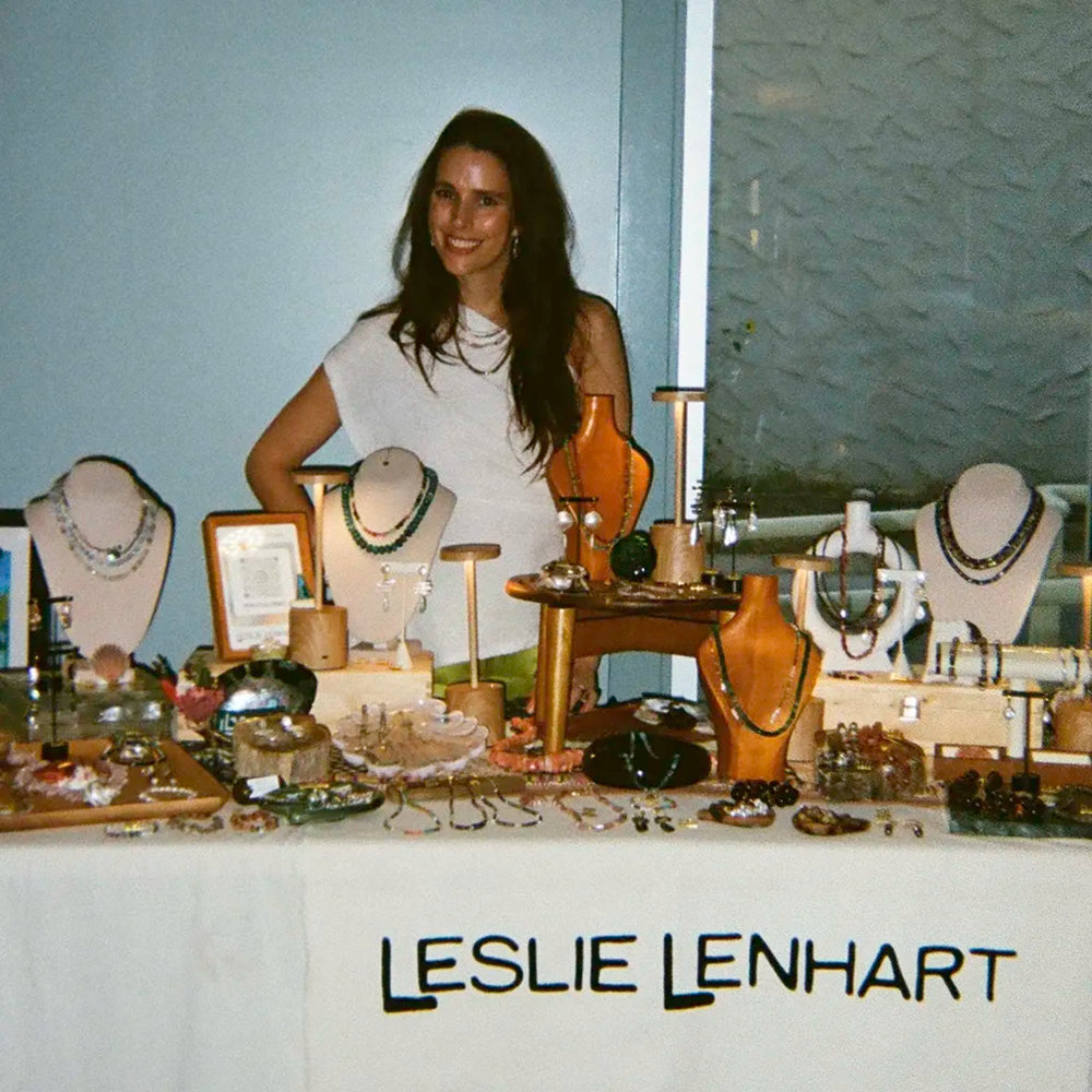 A woman stands behind a display table filled with jewelry on mannequin busts and stands, with the name LESLIE LENHART printed on the front. The scene appears to be an indoor boutique or market-style display with a light blue wall and textured wall panel in the background.
