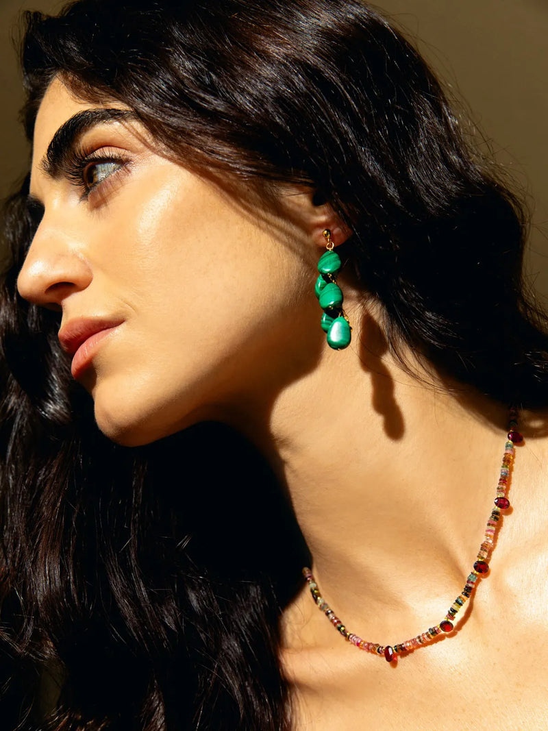 A close-up side profile of a woman with dark hair wearing a green teardrop dangle earring and a colorful beaded necklace against a warm brown background.