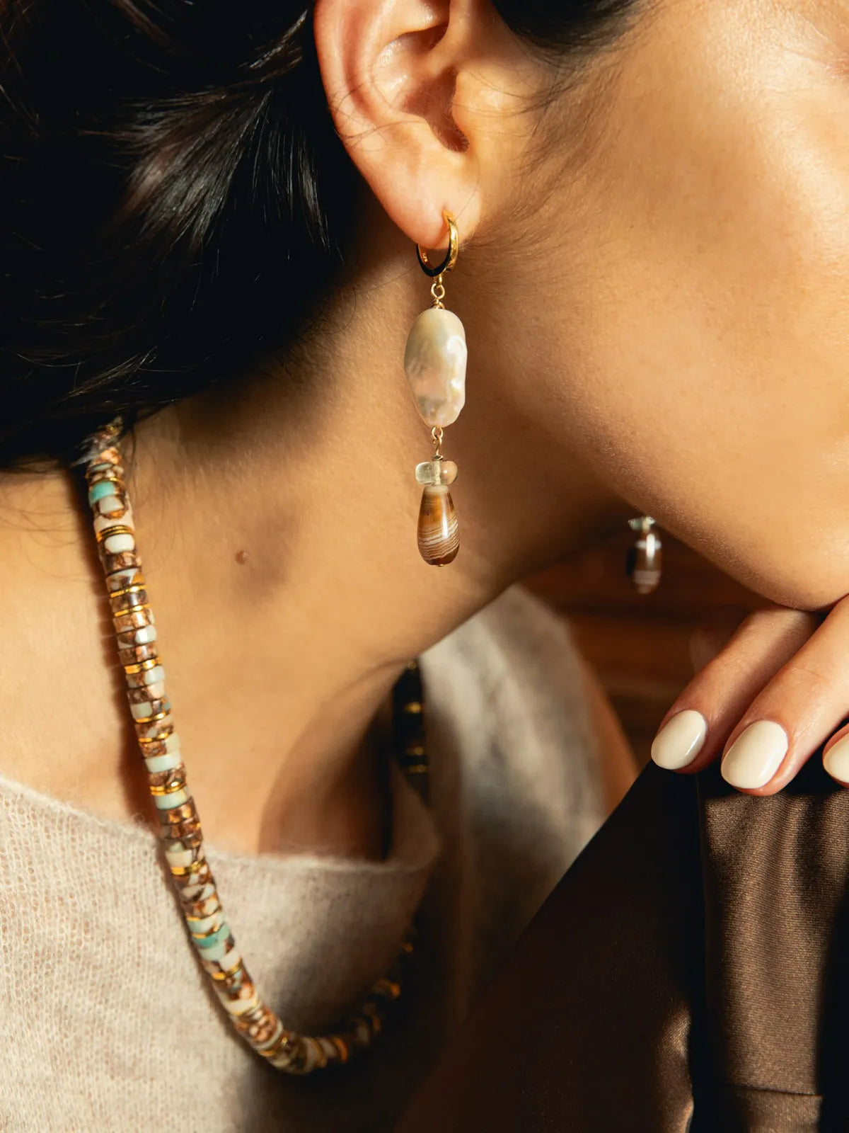 Close-up of a woman wearing a long gold hoop earring with a large irregular cream stone and a smaller amber teardrop drop, paired with a beaded necklace.