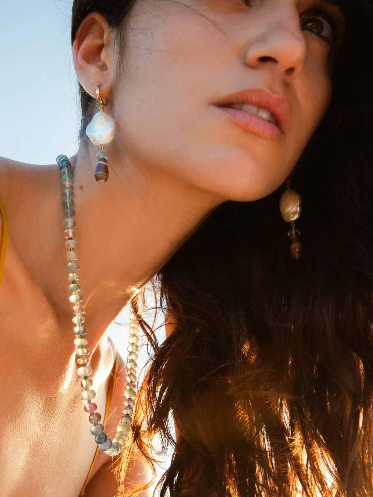 A close-up lifestyle shot of a woman wearing a long beaded necklace and matching drop earrings with large white baroque pearls and small brown beads, set against a bright outdoor sky.