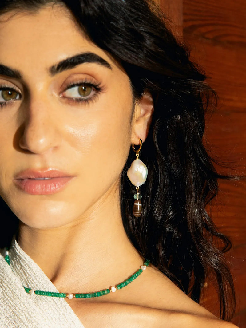 A close-up lifestyle shot of a woman wearing a green beaded necklace and a matching green beaded drop earring with a white pearl and gold details, set against a warm wooden background.