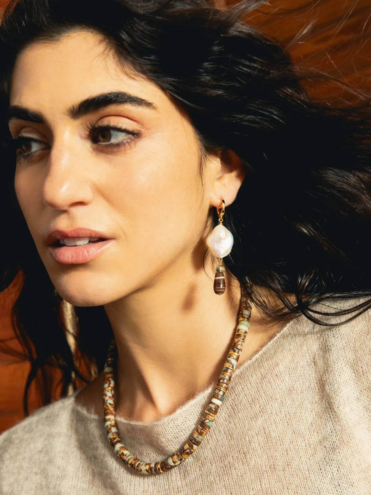 A close-up lifestyle shot of a woman wearing a chunky beaded necklace and a matching drop earring. The jewelry features a mix of earthy brown, gold, and white tones, including a prominent white pearl-like bead in the earring.