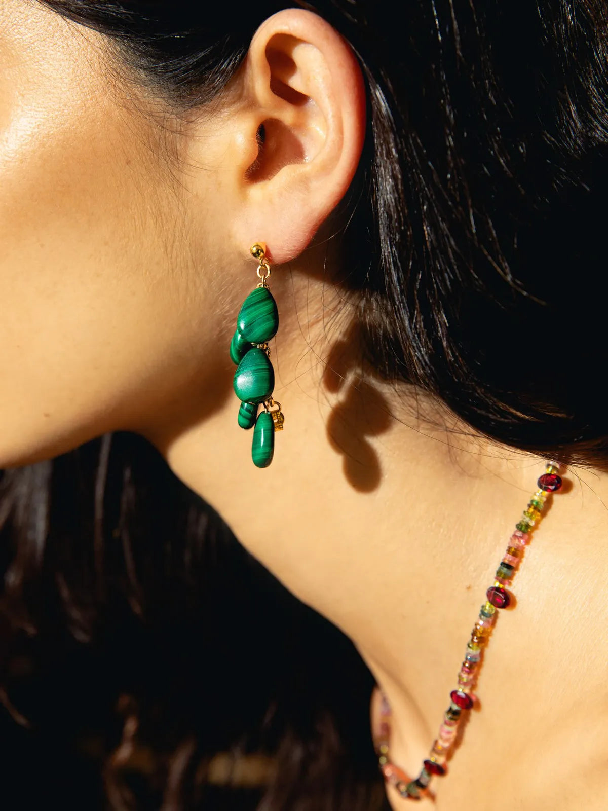 A close-up side profile of a woman wearing a dangling green beaded earring with gold-toned hardware and a matching beaded necklace under warm sunlight.