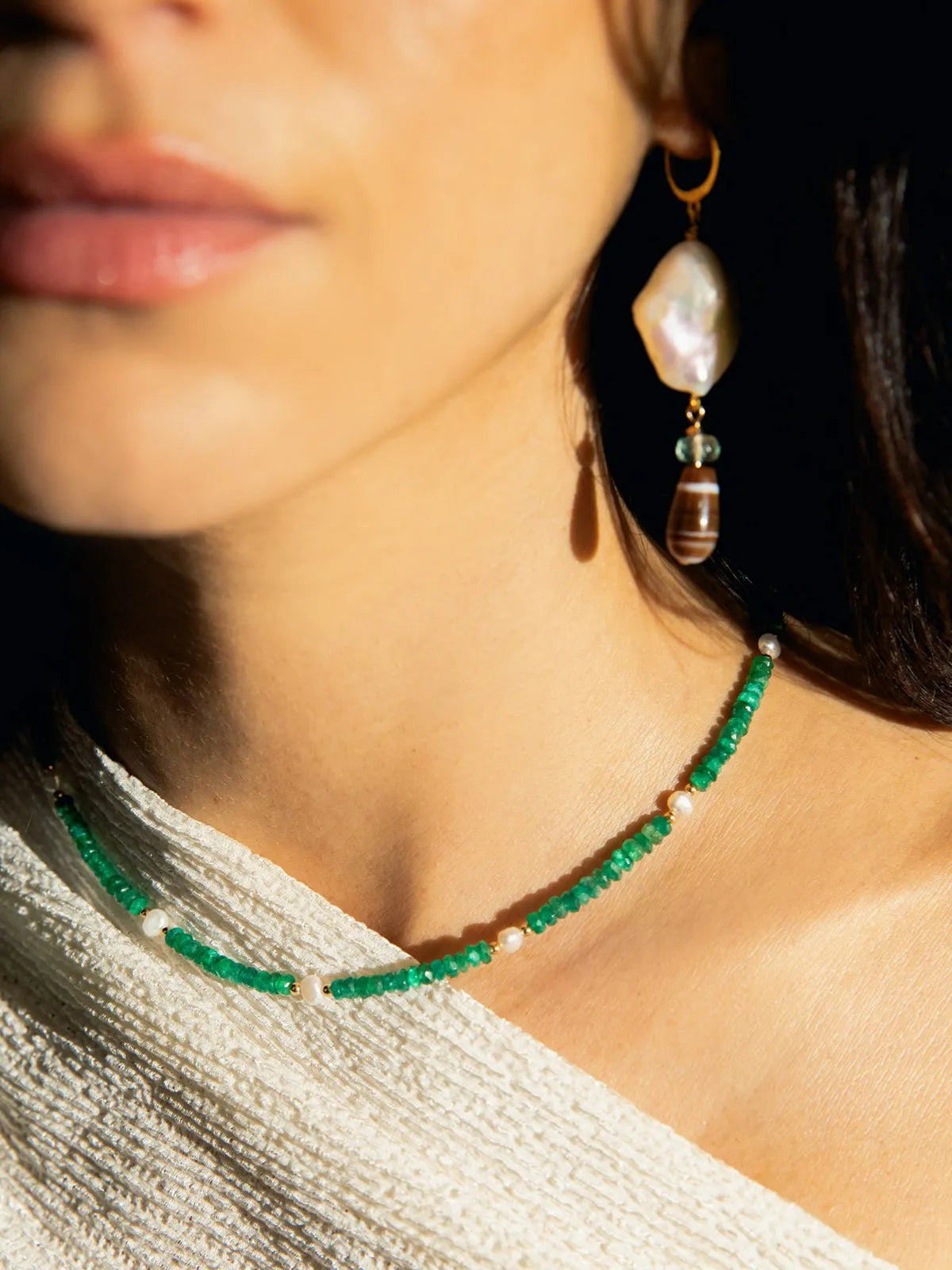 Close-up of a woman wearing a beaded green and white necklace with a large irregular white pearl drop earring, set against a dark background.