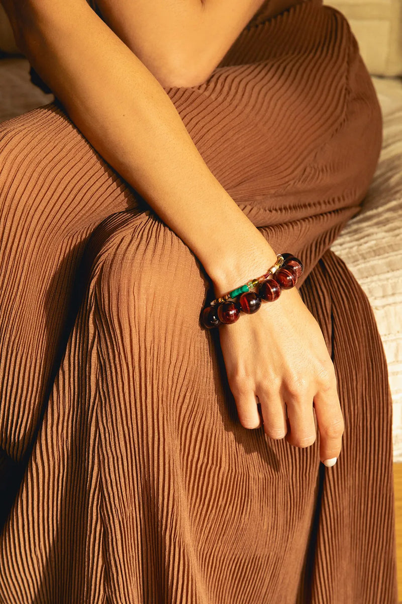 A close-up lifestyle shot of a person wearing a red tiger
s eye beaded bracelet with a small green accent, resting their hand on their lap over a matching pleated brown skirt.