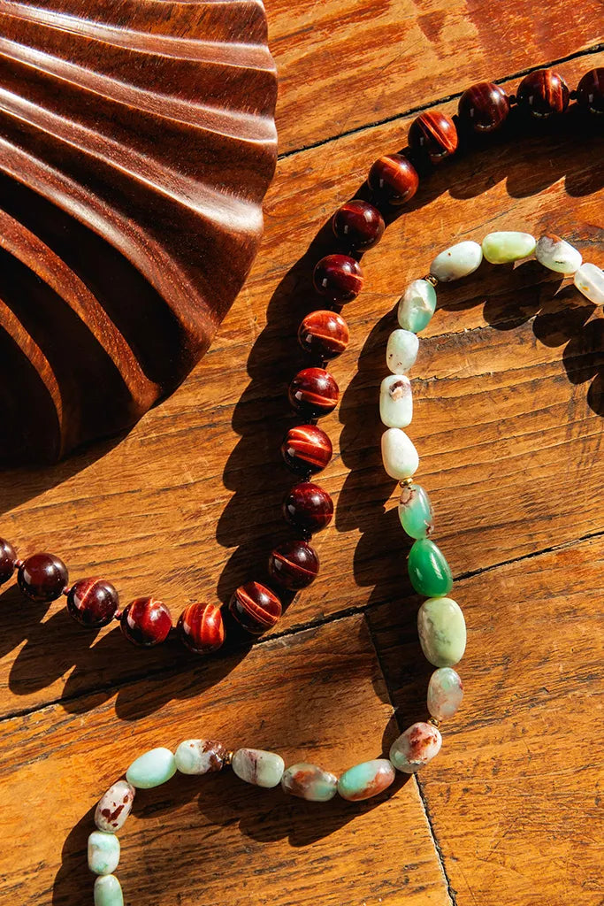 Two beaded necklaces arranged on a warm wooden surface. One necklace features polished dark reddish-brown beads, and the other features light green, white, and marbled beads, with a folded dark wood prop on the left.