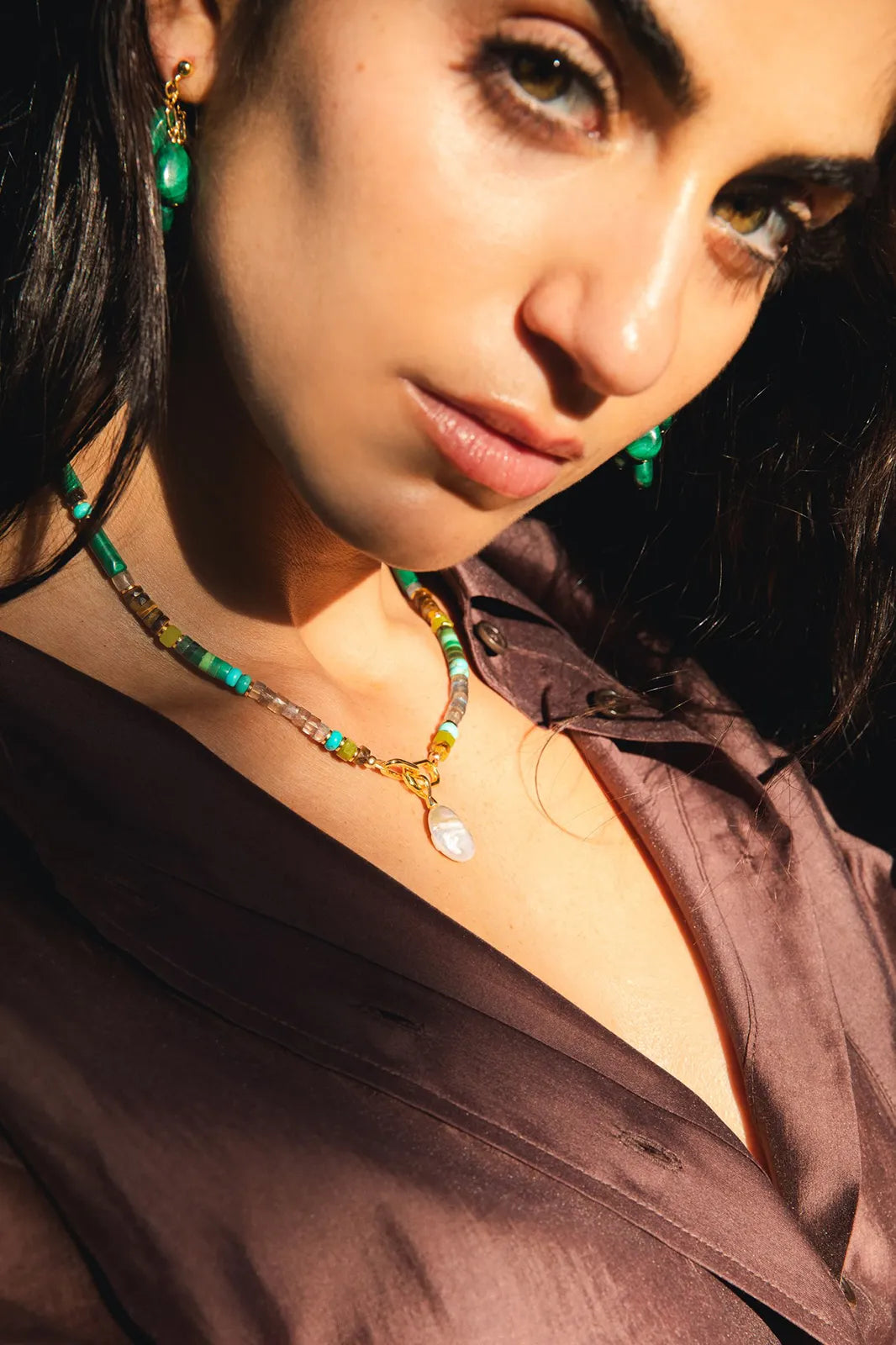 A close-up lifestyle shot of a woman wearing a handmade beaded necklace and matching drop earrings. The necklace features turquoise, green, and brown beads with a small white stone pendant, styled with a brown satin shirt under dramatic warm lighting.