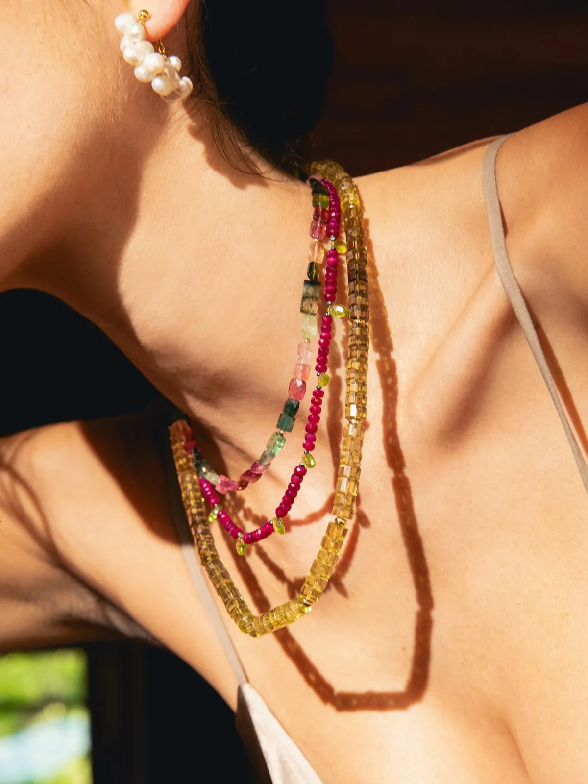 A close-up lifestyle shot of a model wearing a multi-strand beaded necklace with yellow, pink, and green beads, along with a pearl earring, under warm, direct sunlight.
