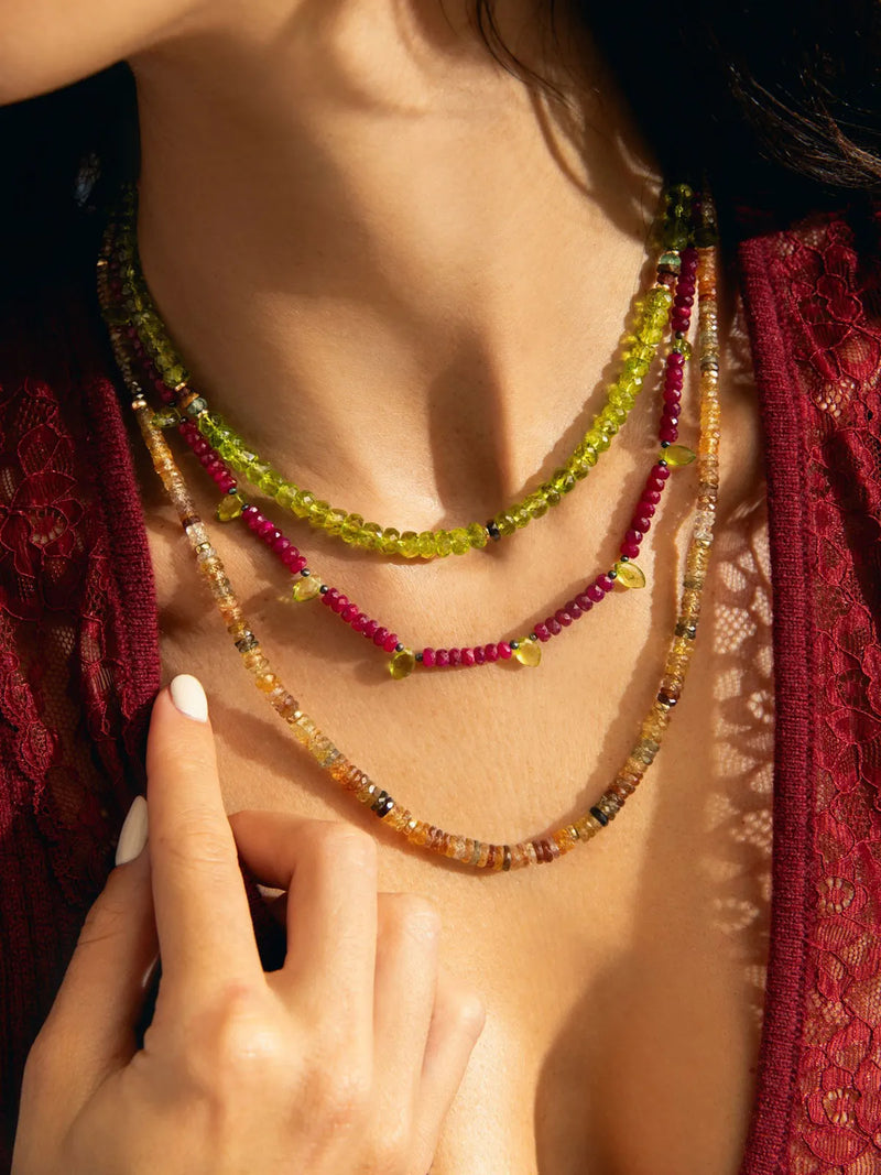 A close-up of a model wearing three layered beaded necklaces. The necklaces feature translucent beads in vibrant shades of yellow-green, deep magenta pink, and warm amber-brown.