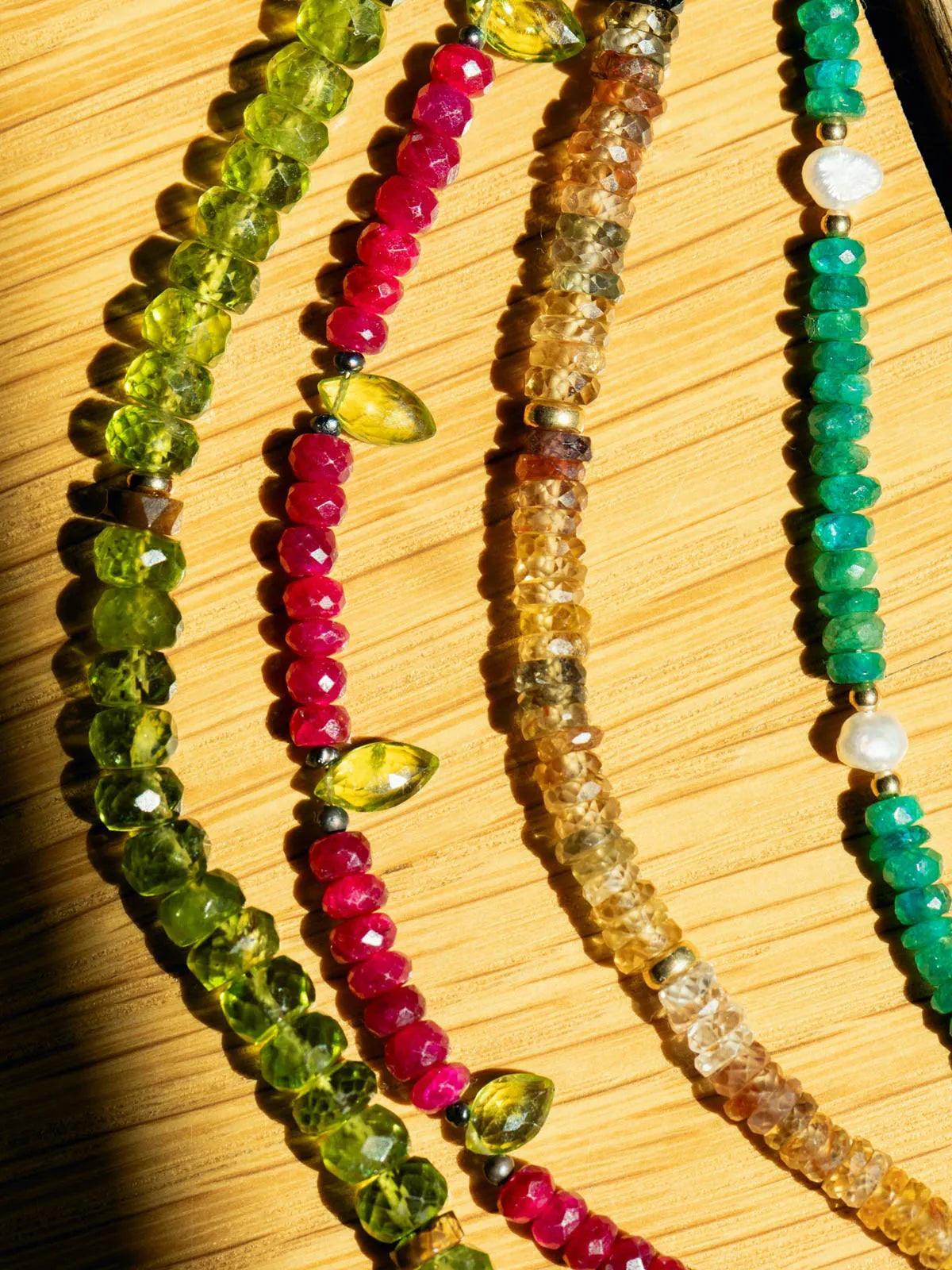 A close-up of several beaded strands laid diagonally on a light wood surface. The beads include green, pink, amber-brown, and turquoise tones, with faceted round and teardrop-shaped stones separated by small metallic spacer beads.