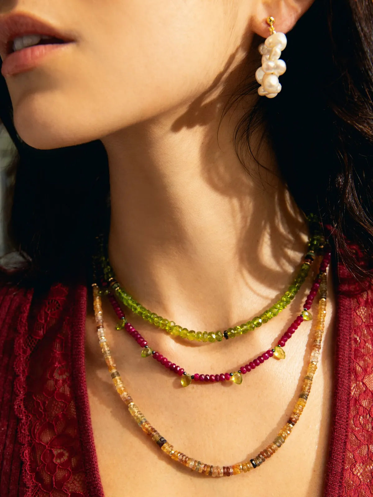 Close-up of a woman wearing layered beaded necklaces and a dangling pearl earring. The necklaces feature green, magenta, and amber-toned beads, with the woman wearing a dark red lace top.