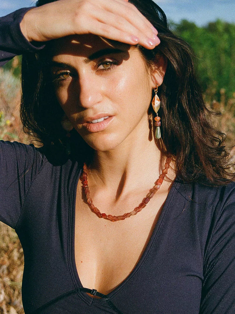 Woman with dark hair and earrings, wearing a necklace, outdoors.