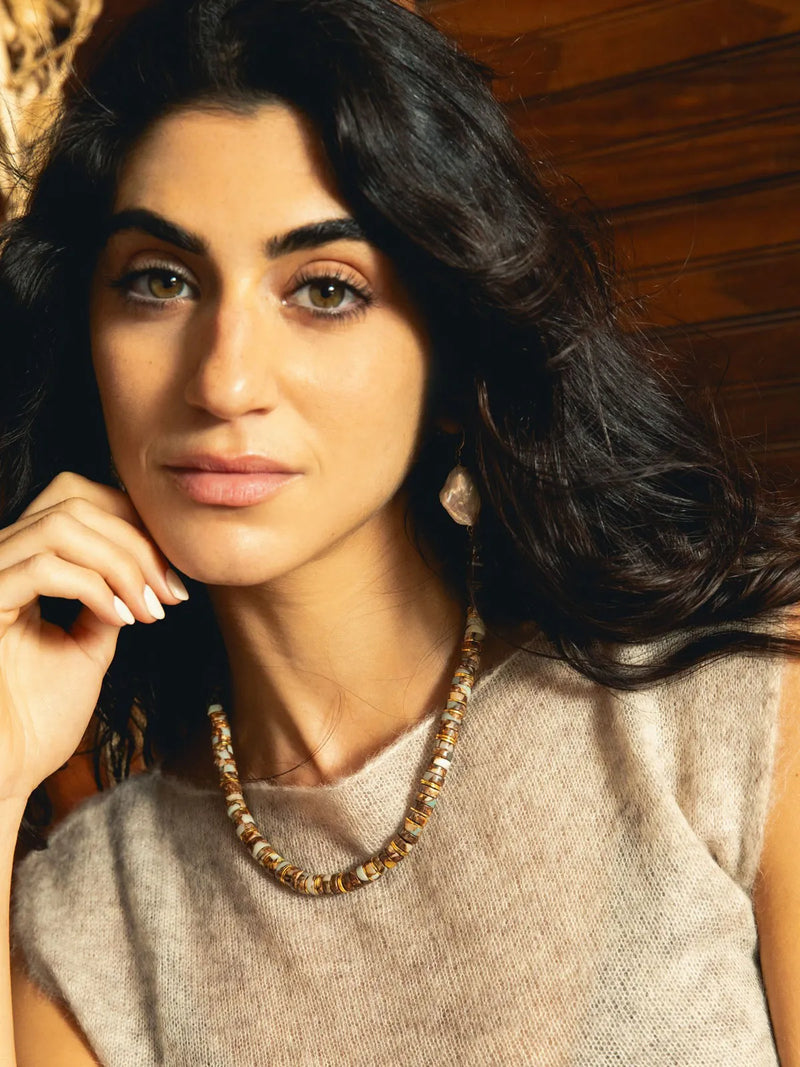 A close-up portrait of a woman with dark hair wearing a chunky gold and brown beaded necklace and matching earrings, set against a warm wooden background.