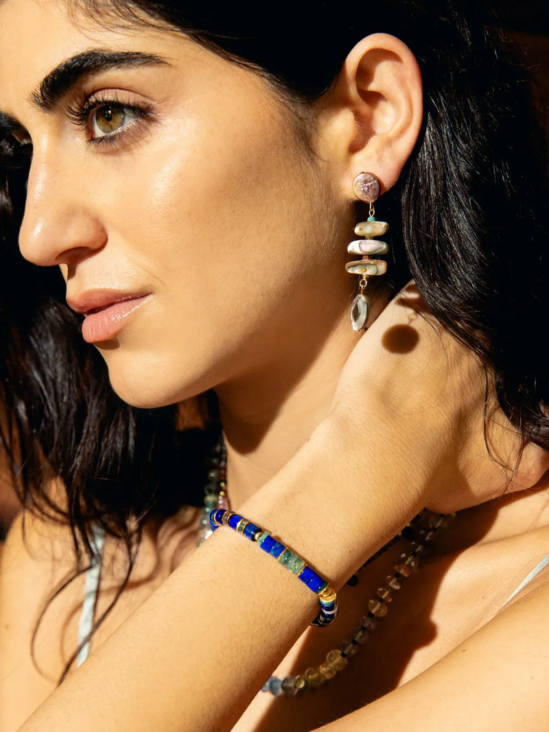 A close-up lifestyle shot of a woman wearing bohemian-style jewelry, featuring a long dangle earring with stacked natural stones and a matching beaded bracelet on her wrist.