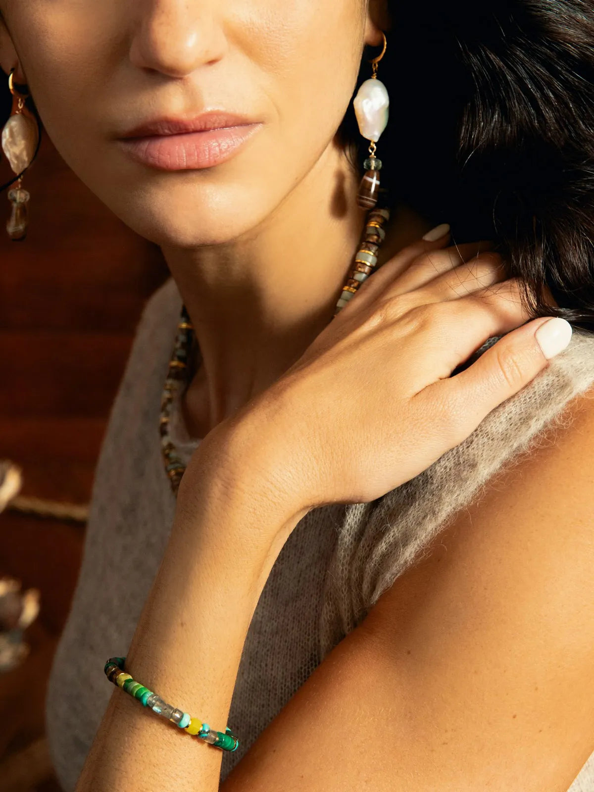 A close-up lifestyle shot of a woman wearing a colorful beaded bracelet, matching beaded necklace, and large white dangle earrings against a warm, dark background.