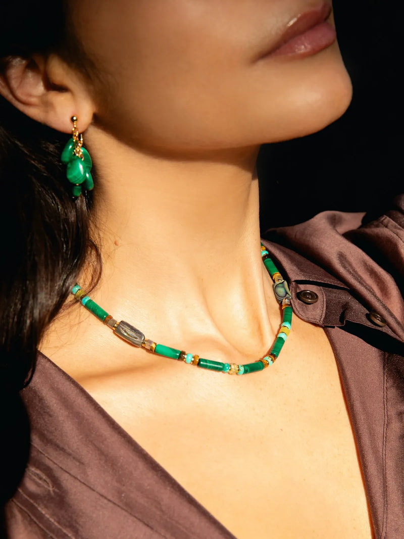 A close-up lifestyle shot of a model wearing a beaded necklace and matching drop earrings. The jewelry features vibrant green beads, small gold spacer beads, and polished gray stone accents.