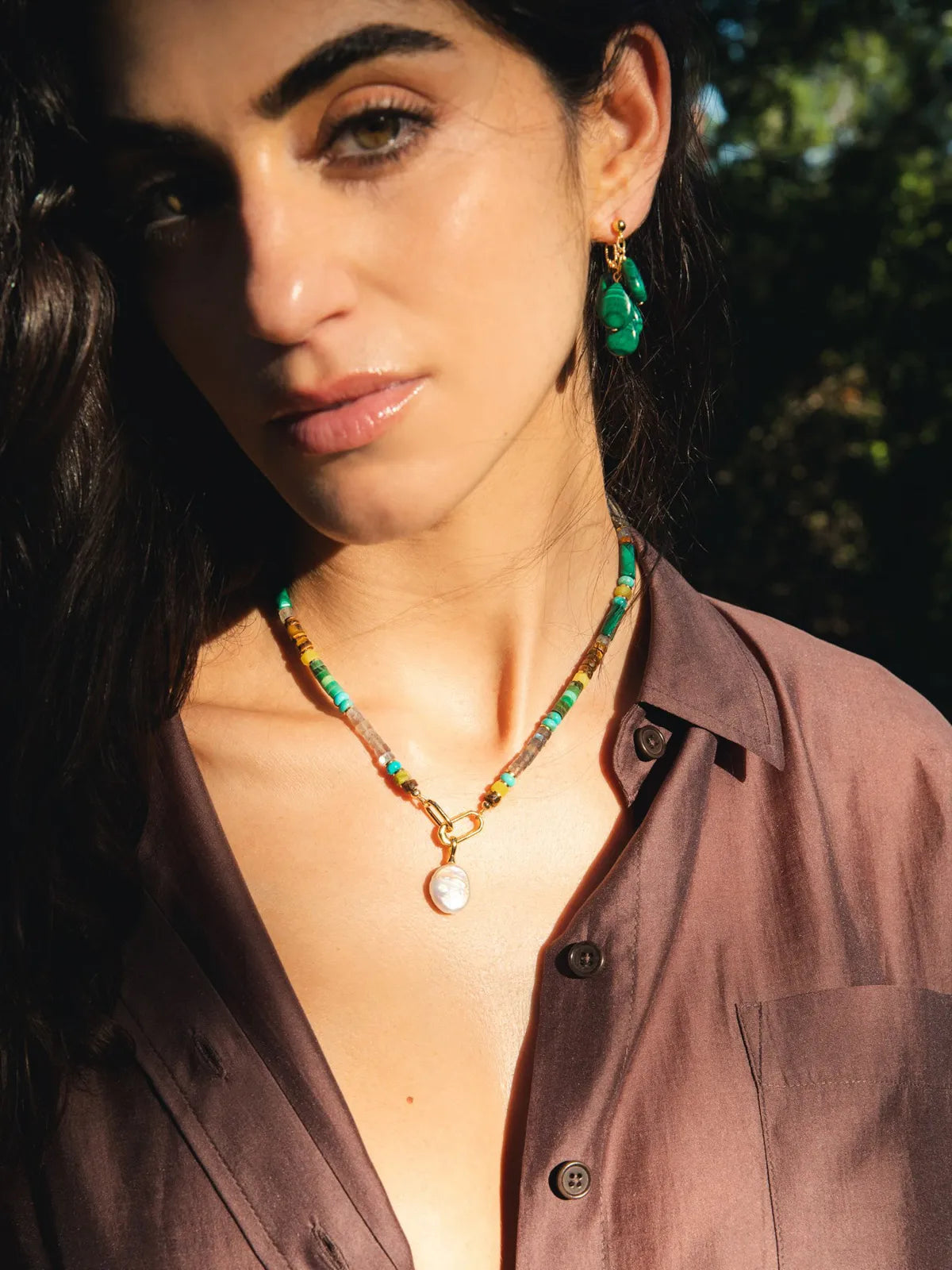 A woman wearing a colorful beaded necklace with a white pearl pendant and matching green drop earrings, photographed outdoors in warm sunlight.