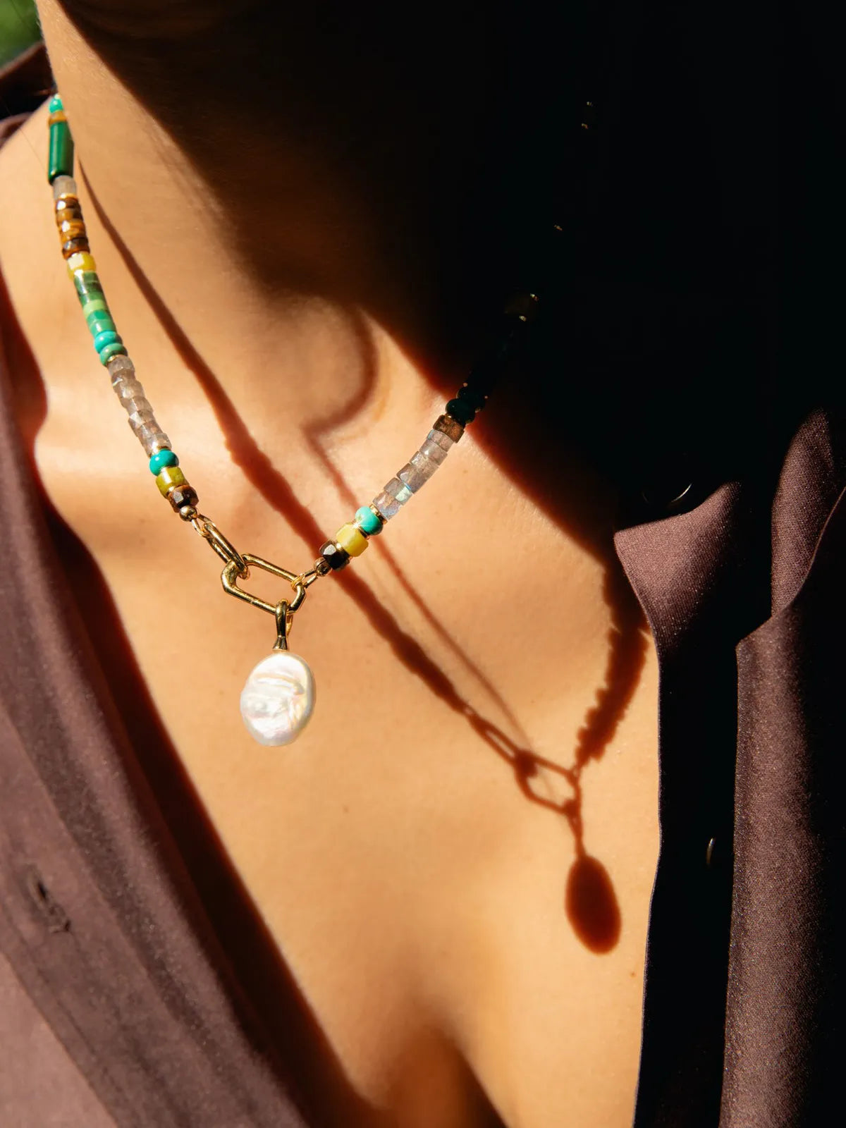 A close-up of a person wearing a beaded necklace with a white oval pendant, styled with a dark brown top under dramatic warm sunlight and shadows.
