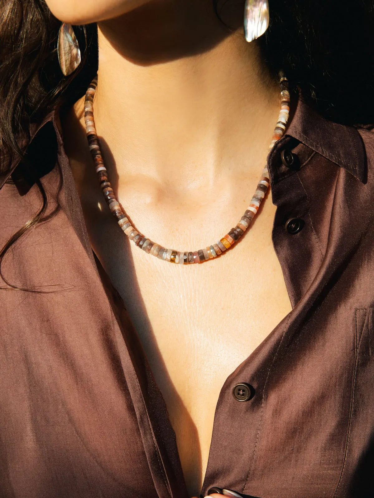 A close-up of a woman wearing a chunky beaded necklace made of terracotta, brown, and cream stone-like beads, paired with a brown button-up shirt and silver earrings.