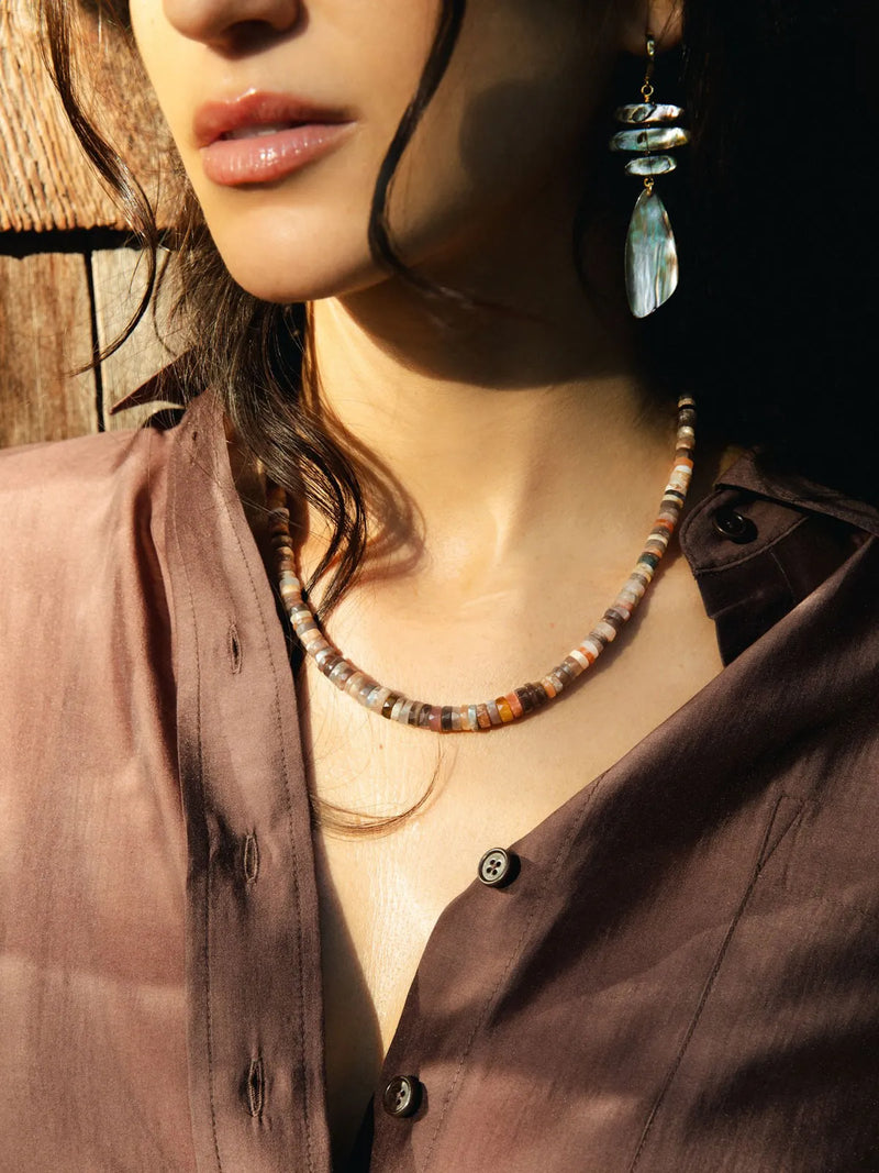 A close-up lifestyle shot of a woman wearing a beaded necklace and matching drop earring. The necklace features a mix of earthy, multi-toned beads in brown, beige, and orange.