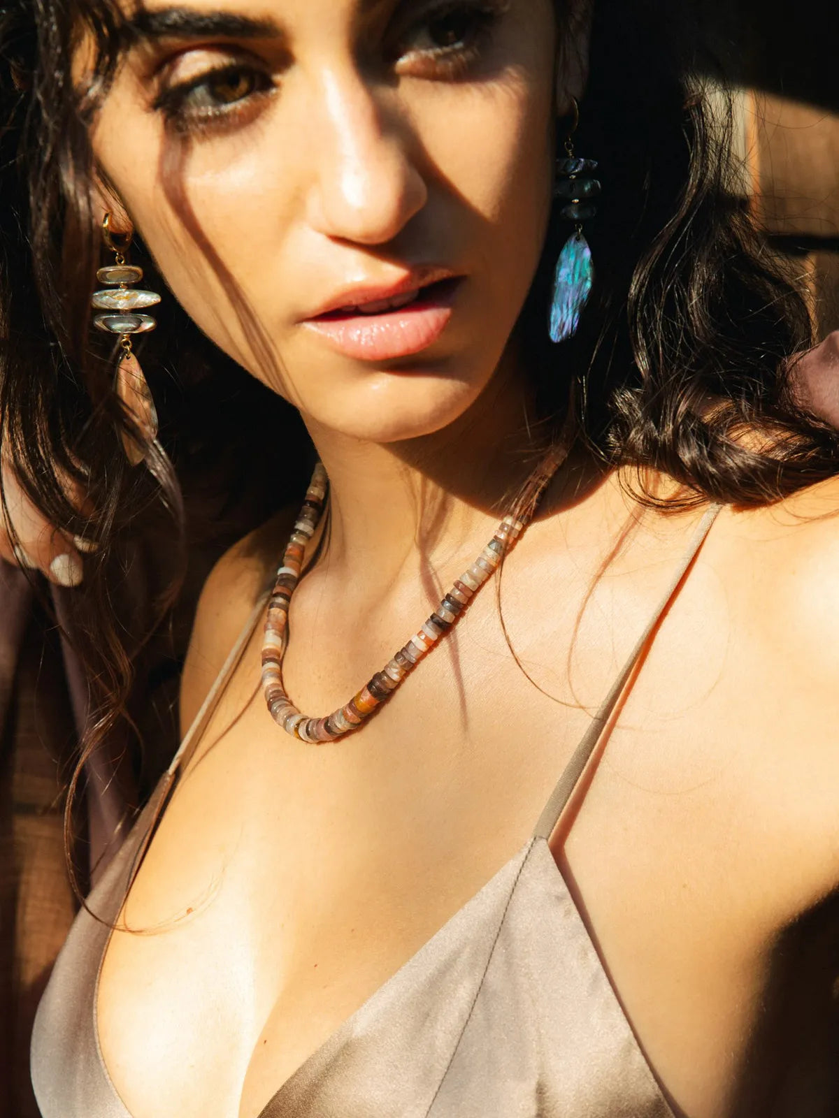 A close-up lifestyle shot of a woman wearing a beaded necklace and matching drop earrings. The necklace features earthy brown, beige, and white beads, while the earrings have blue and white accents.