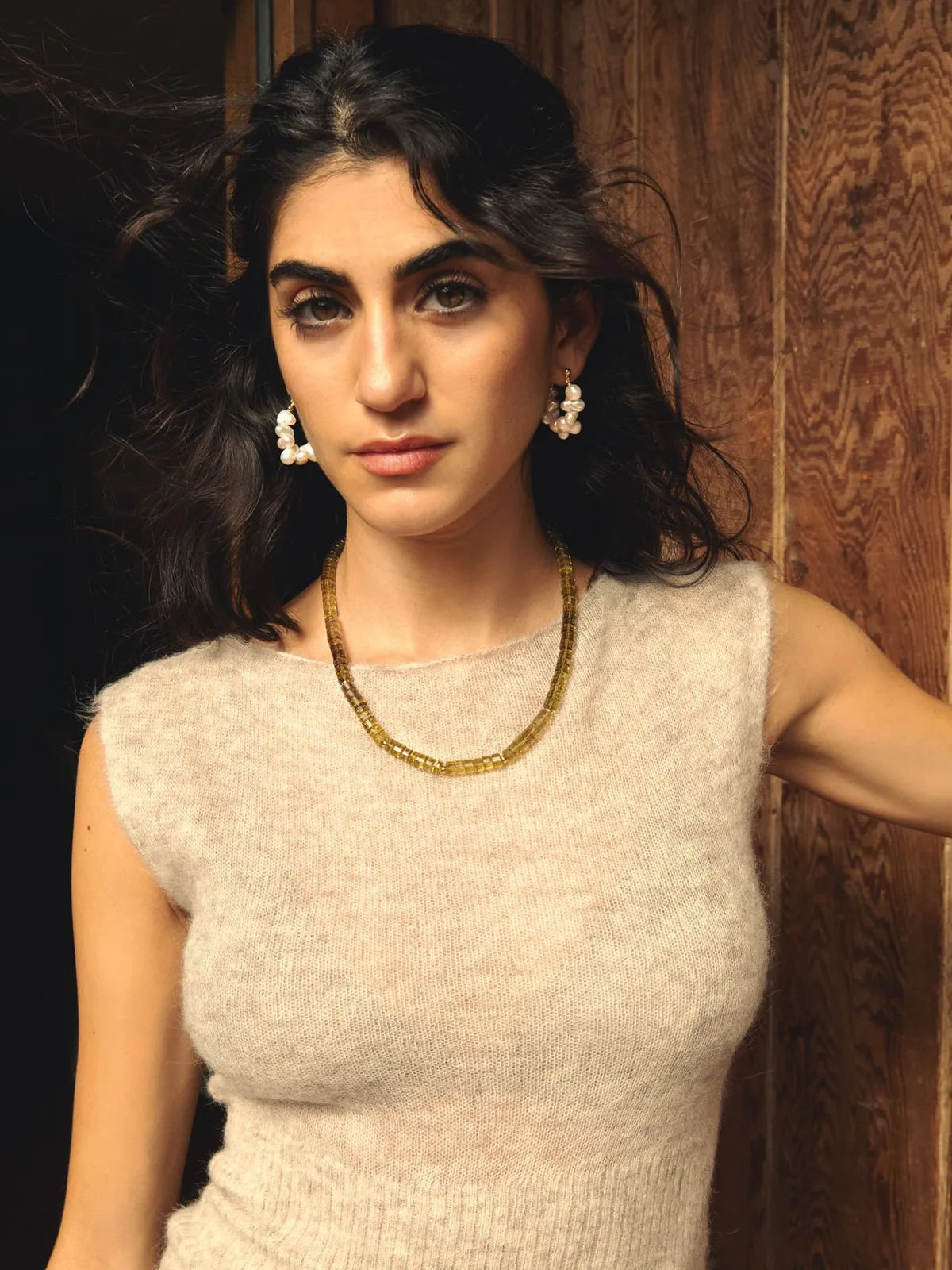 A woman with dark hair wearing a textured cream knit sleeveless top, a gold chain necklace, and pearl drop earrings, posing against a rustic wooden wall.