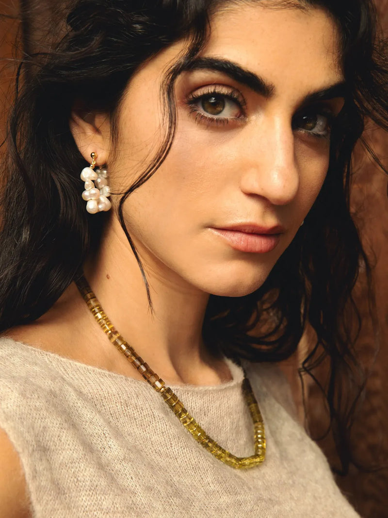 A close-up portrait of a woman wearing a gold-toned beaded necklace and matching drop earrings with white pearl-like beads, against a warm brown background.
