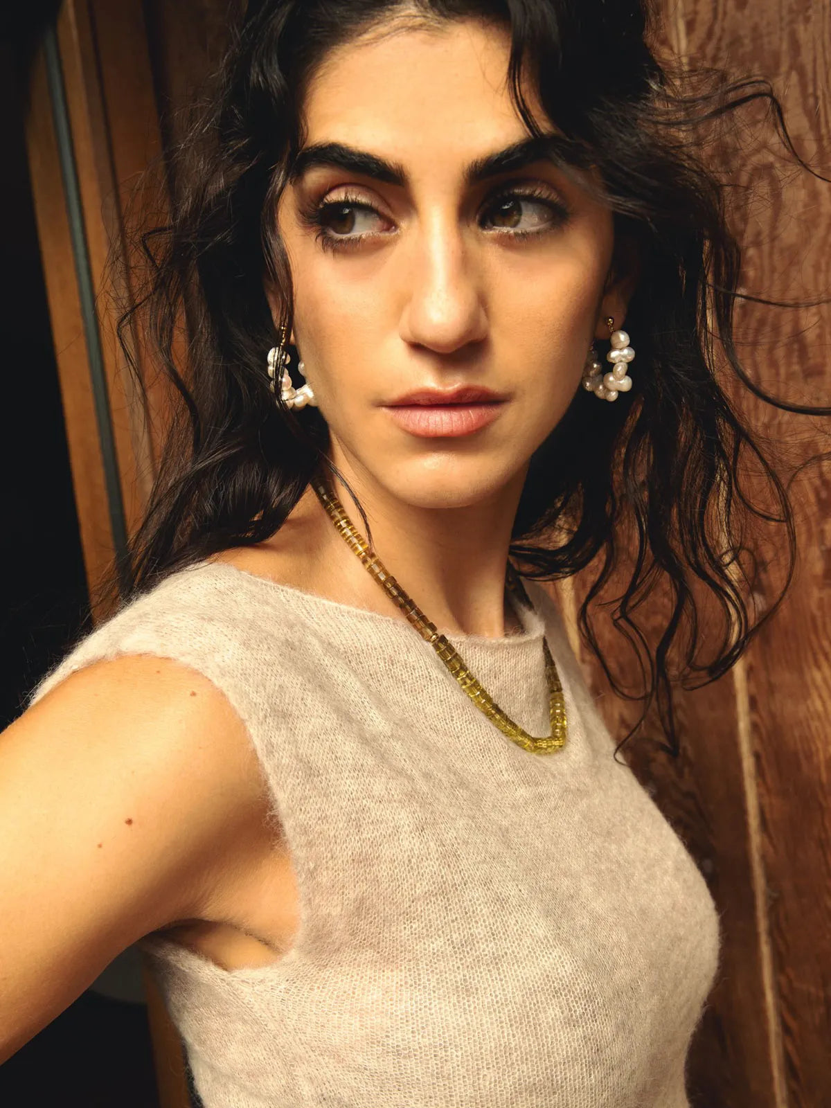 A close-up portrait of a woman with dark wavy hair wearing a light beige knit top, a gold chain necklace, and pearl-like drop earrings, set against a dark wood background.