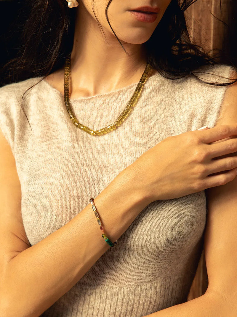 A close-up lifestyle shot of a woman wearing a gold beaded necklace and a multi-colored beaded bracelet, paired with a light beige knit top.
