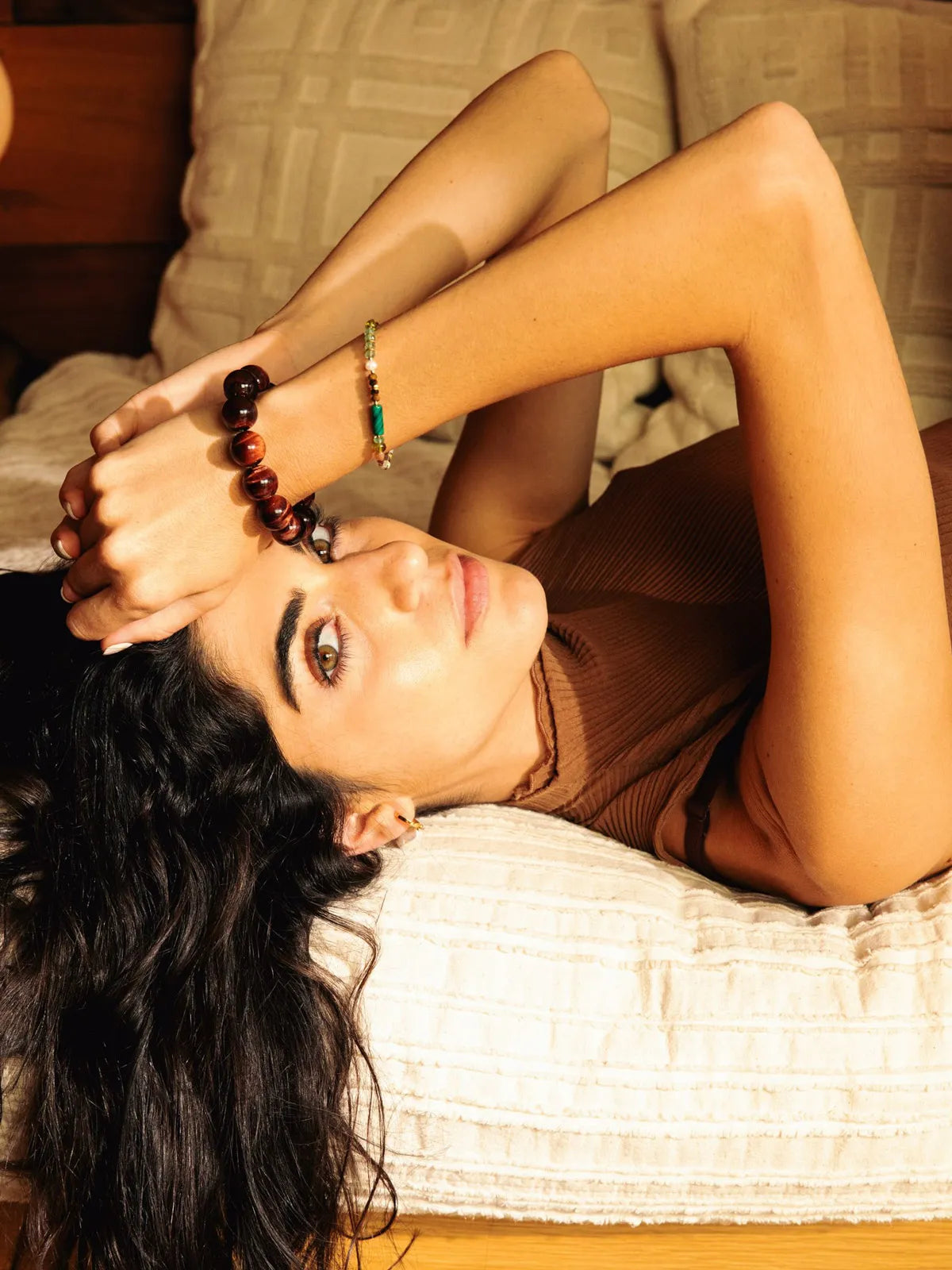 A woman lying on a bed wearing a dark brown beaded bracelet and a green beaded bracelet, with warm golden lighting.