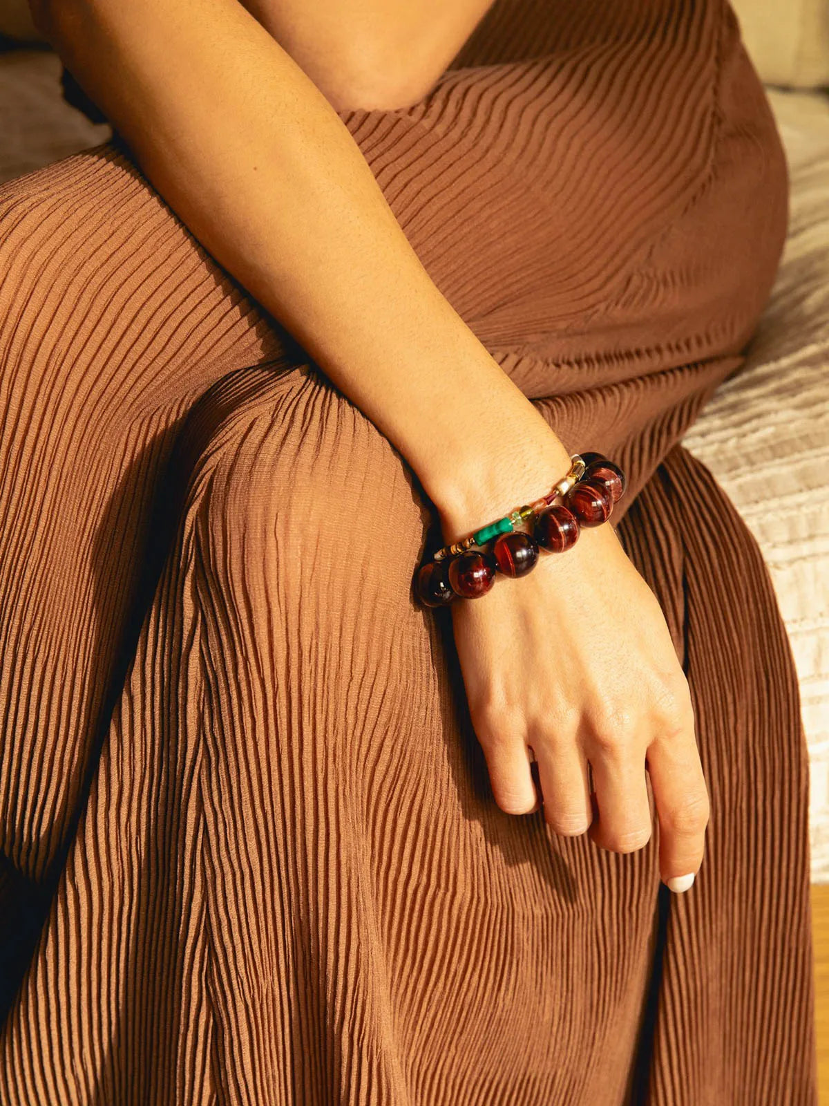 A close-up of a person's hand resting on their lap, wearing a dark reddish-brown beaded bracelet with a small green and gold accent. The person is wearing a matching pleated brown skirt in warm, golden lighting.