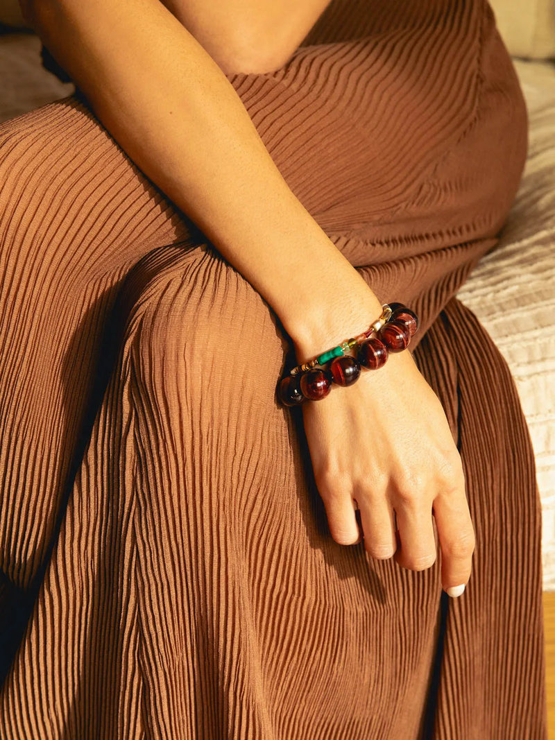 A close-up of a person's hand resting on their lap, wearing a dark reddish-brown beaded bracelet with a small green and gold accent. The person is wearing a matching pleated brown skirt in warm, golden lighting.