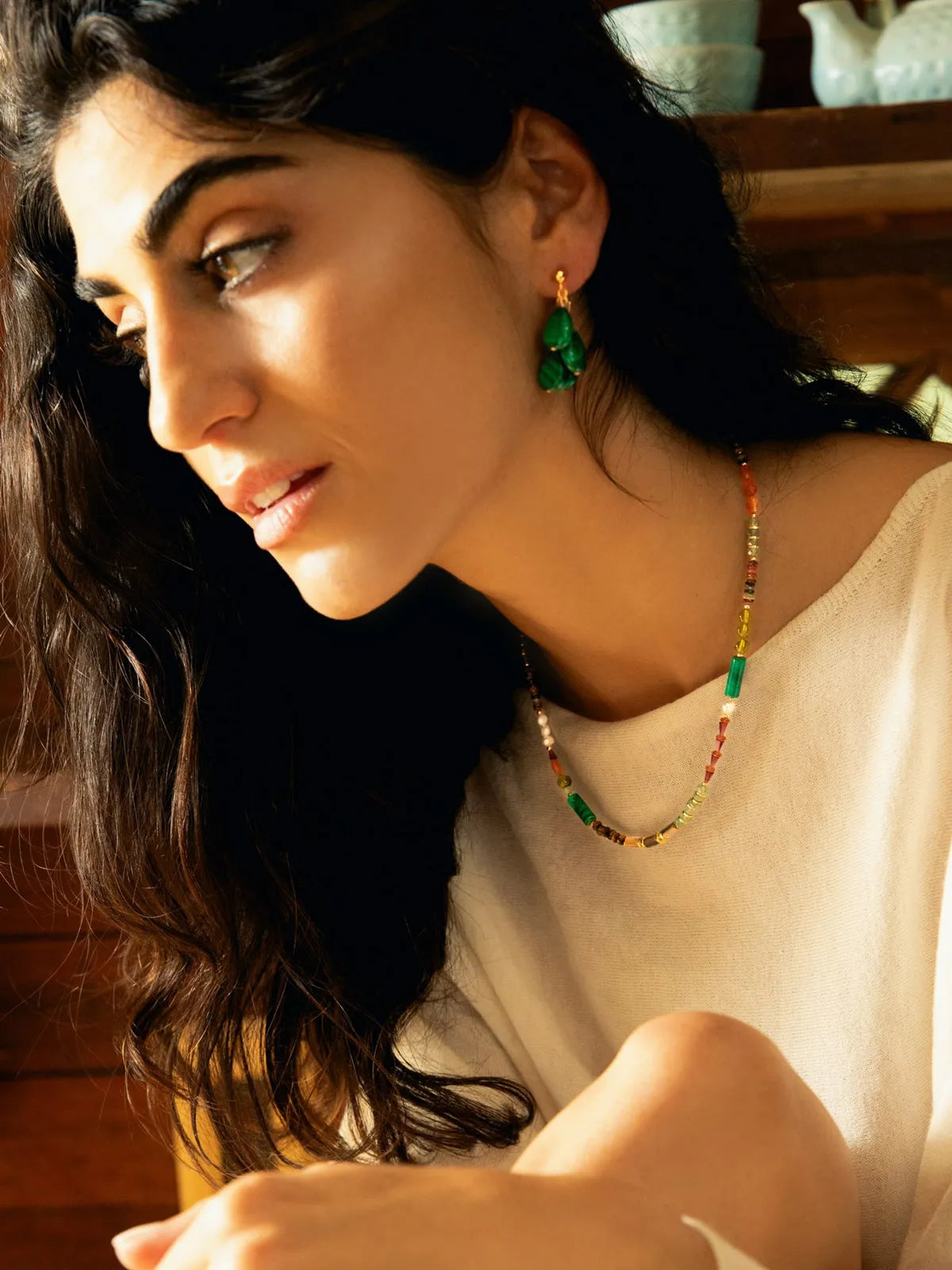 A woman wearing a colorful beaded necklace and matching green drop earrings, shown in a warm, sunlit indoor setting.