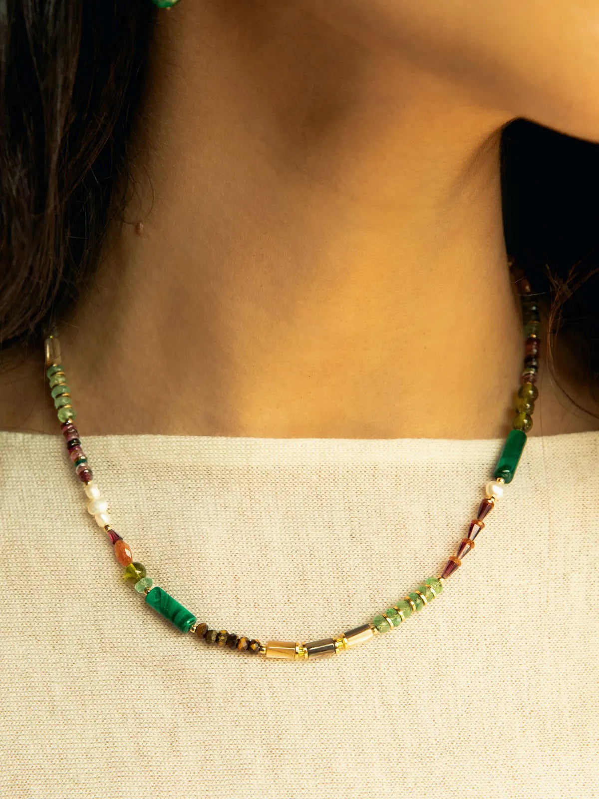A close-up of a person's neck and upper chest wearing a delicate beaded necklace. The necklace features green cylindrical beads, small white pearls, and translucent amber-colored beads separated by small gold-toned spacer beads.