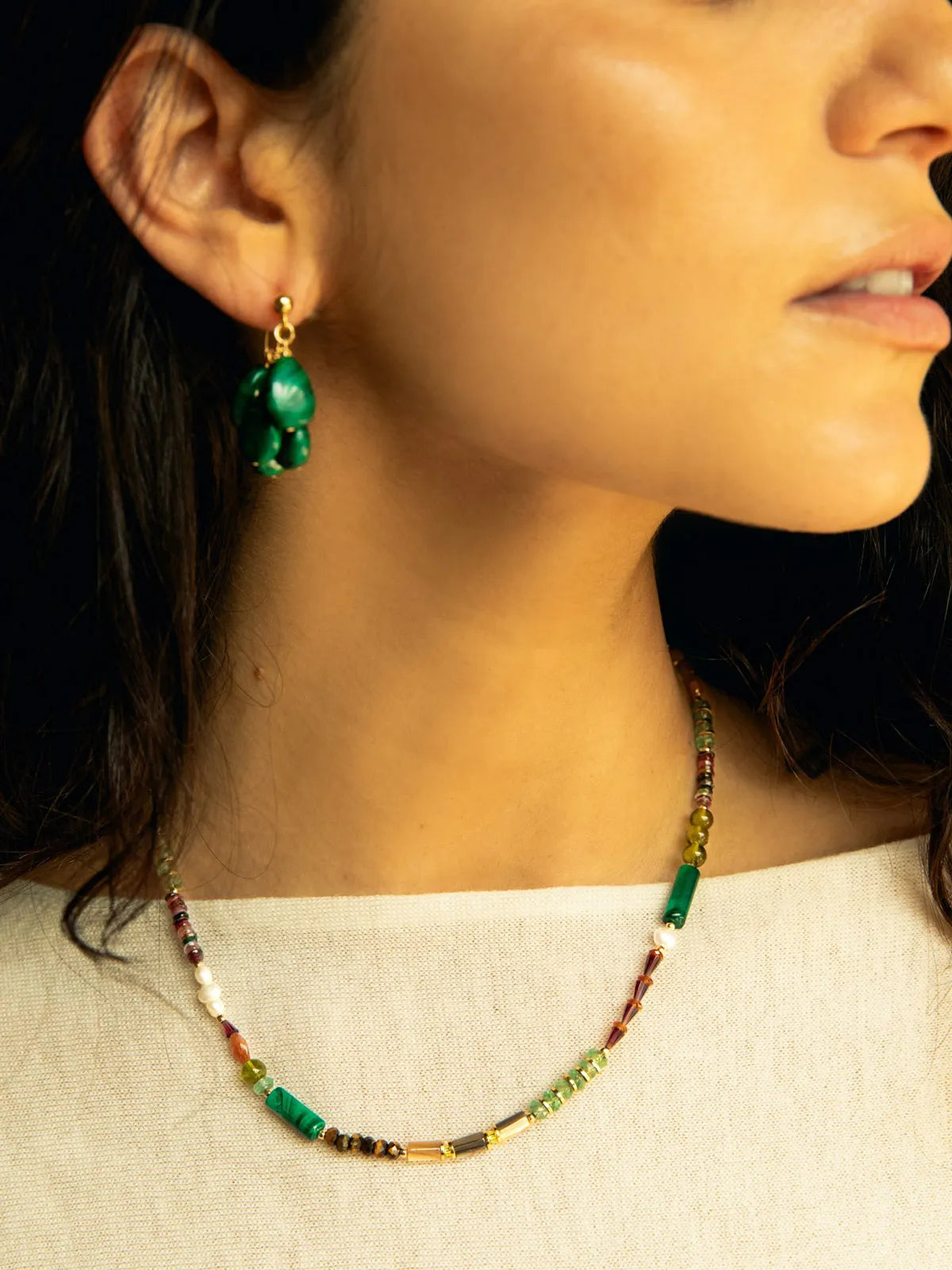 Close-up of a woman wearing a beaded necklace and matching drop earring. The jewelry features green beads, gold-toned metal, and small red and white accents against a dark background.