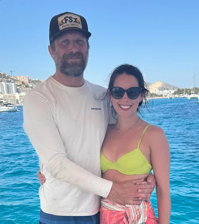 Man and woman standing together by a body of water with a clear blue sky.