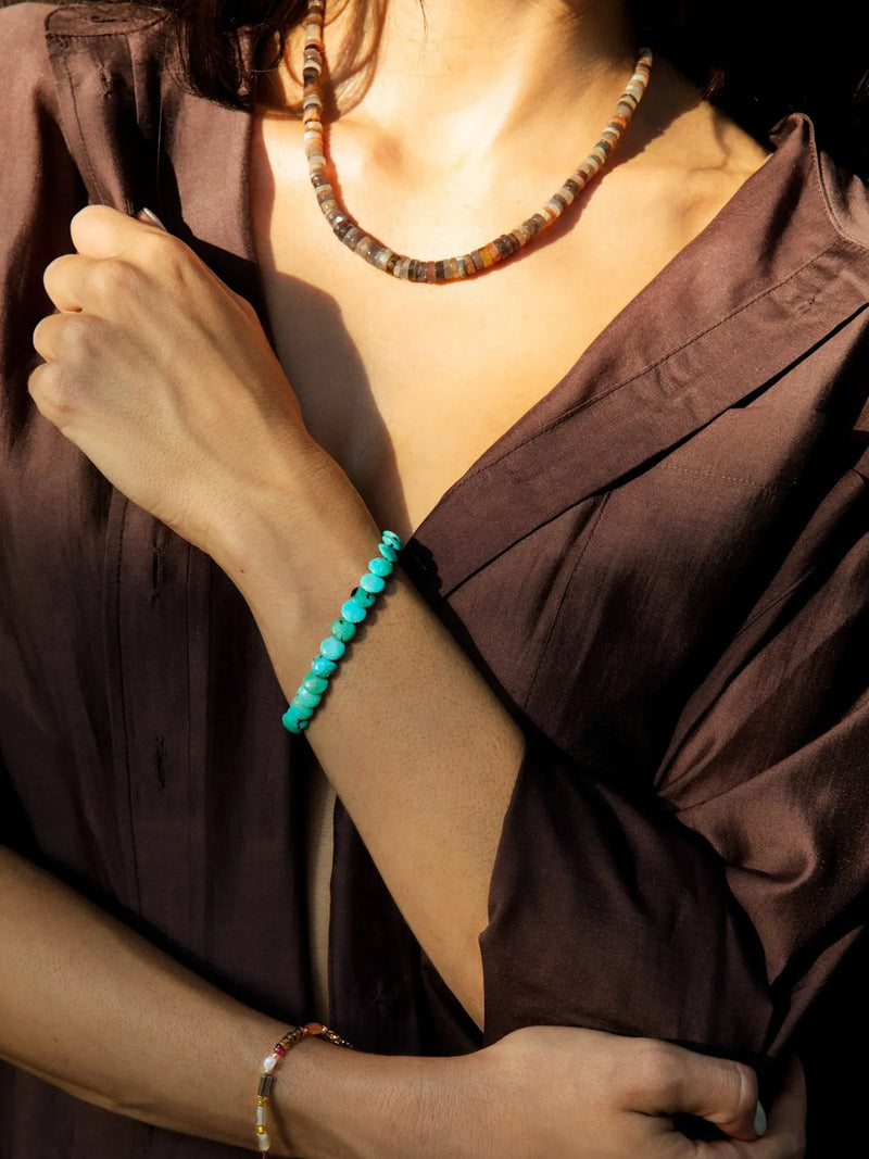 A close-up lifestyle shot of a woman wearing a vibrant turquoise beaded bracelet on her wrist, a multi-colored beaded necklace, and a delicate gold-toned bracelet, styled with a brown satin blouse.