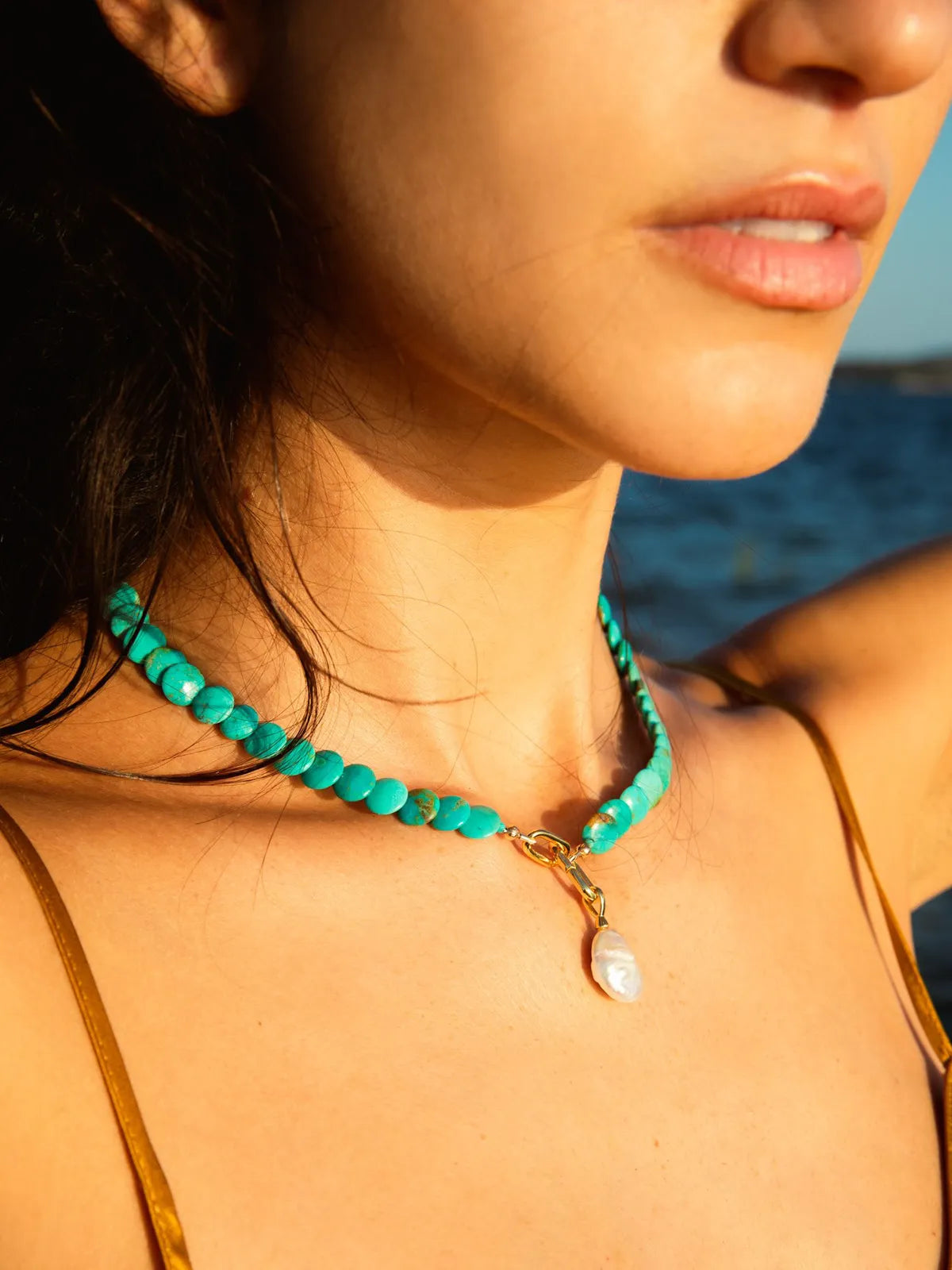 A close-up lifestyle shot of a woman wearing a turquoise beaded choker necklace with a white pearl pendant and gold clasp, set against a warm outdoor background.