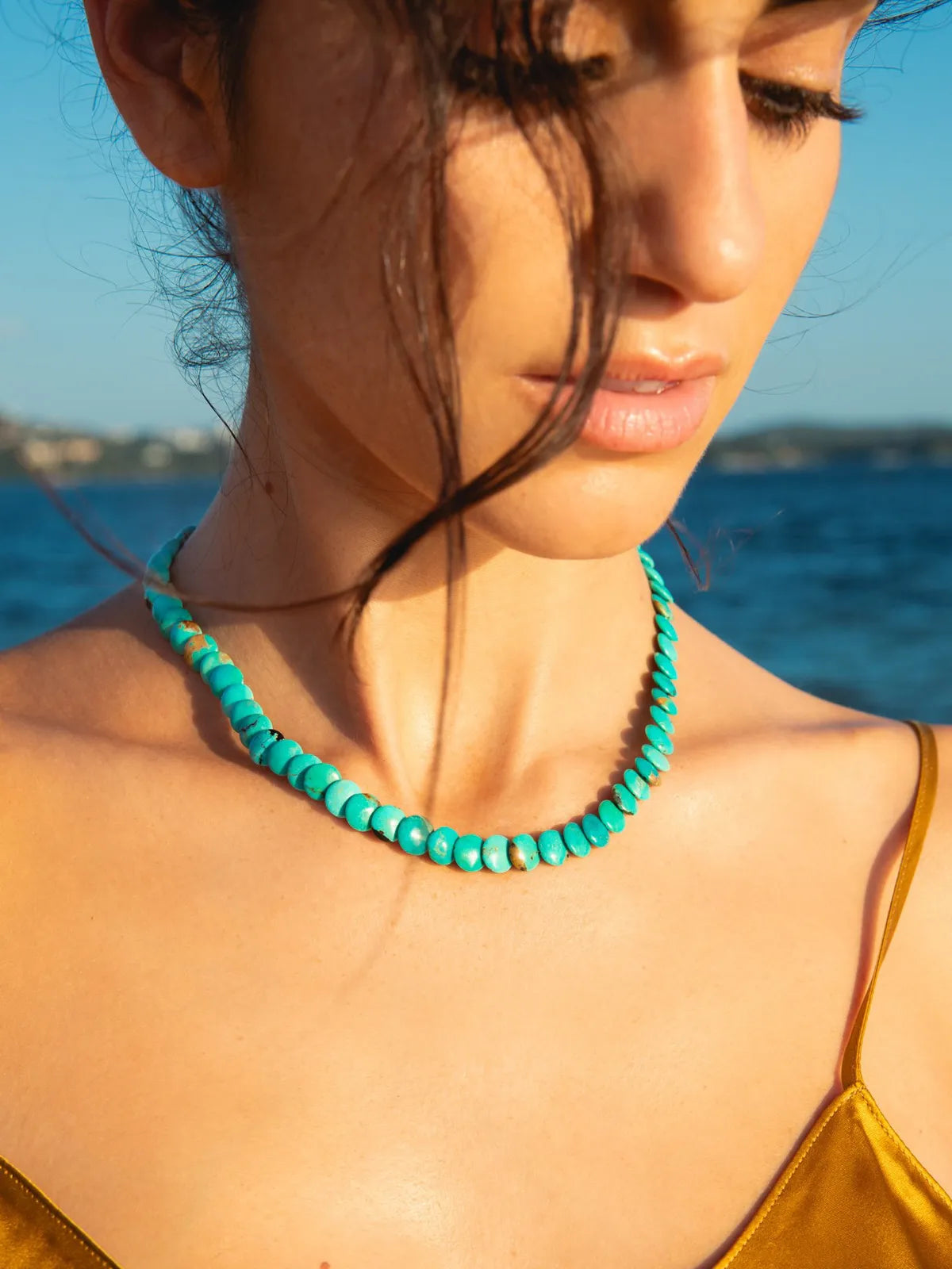 A close-up lifestyle shot of a woman wearing a turquoise beaded necklace, with the ocean and sky blurred in the background.