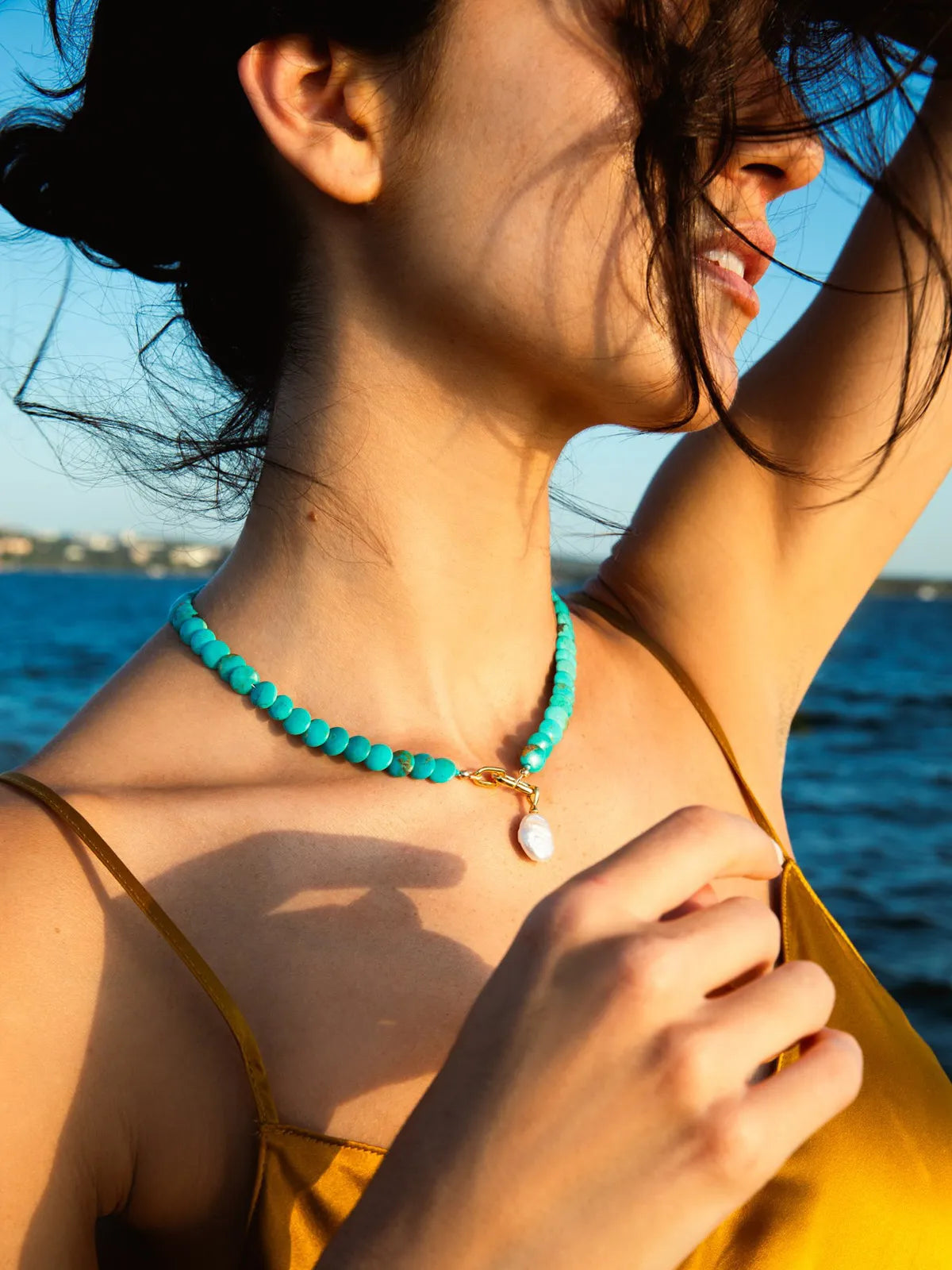A woman outdoors by the sea wearing a turquoise beaded necklace with a small white shell pendant and gold clasp, paired with a yellow top.