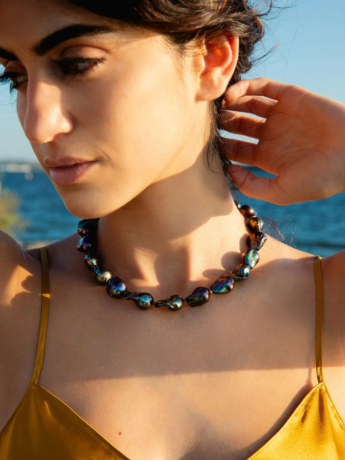 A close-up lifestyle shot of a woman outdoors by the sea, wearing a dark iridescent beaded necklace with baroque-style beads.