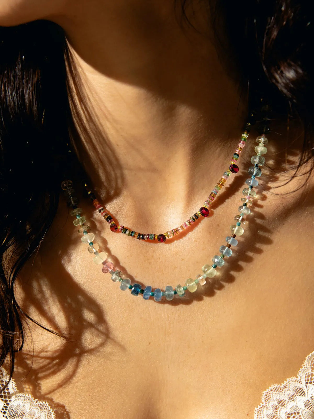 A close-up of a model wearing two layered beaded necklaces. The top necklace features small multicolored beads, and the bottom necklace features larger faceted beads in shades of blue, green, pink, and yellow.