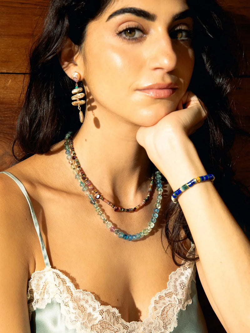 A woman with dark hair resting her chin on her hand, wearing a layered beaded necklace, matching beaded earrings, and a beaded bracelet against a warm wooden background.