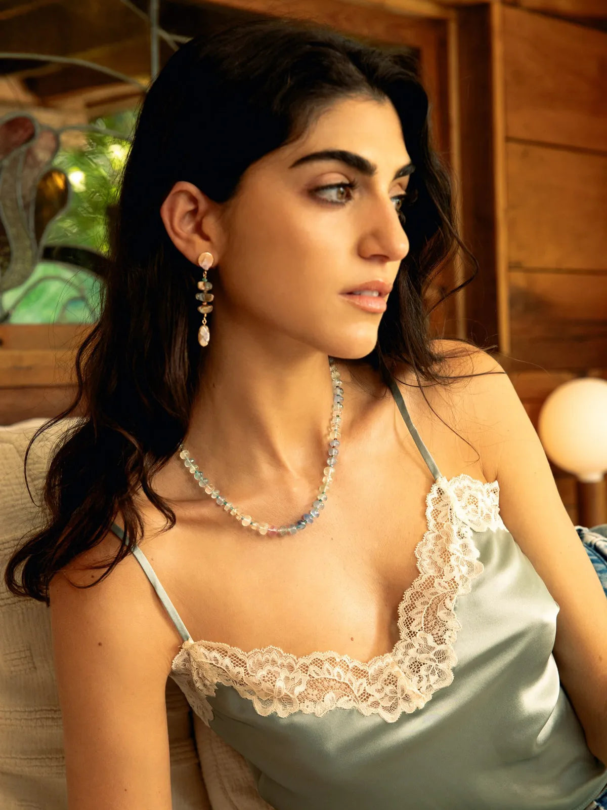A woman wearing a light sage green satin camisole with white lace trim, styled with a matching beaded necklace and dangling earrings in a warm, rustic indoor setting.