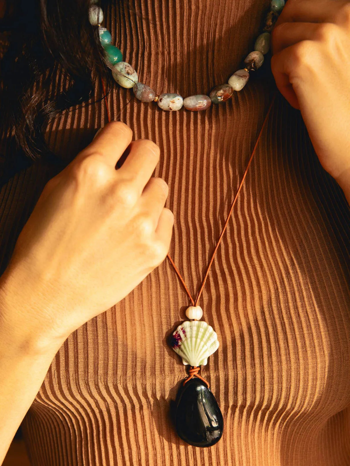 A close-up lifestyle shot of a model wearing a brown ribbed knit top, showcasing a long brown cord necklace with a shell and black stone pendant, alongside a shorter beaded necklace.