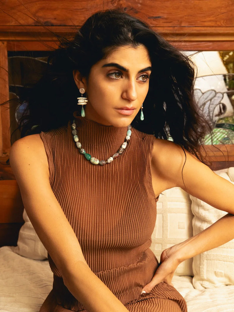 A woman with long dark hair wearing a brown ribbed sleeveless turtleneck dress, a matching brown pleated top, and turquoise and white beaded jewelry, sitting on a bed with a wooden headboard.