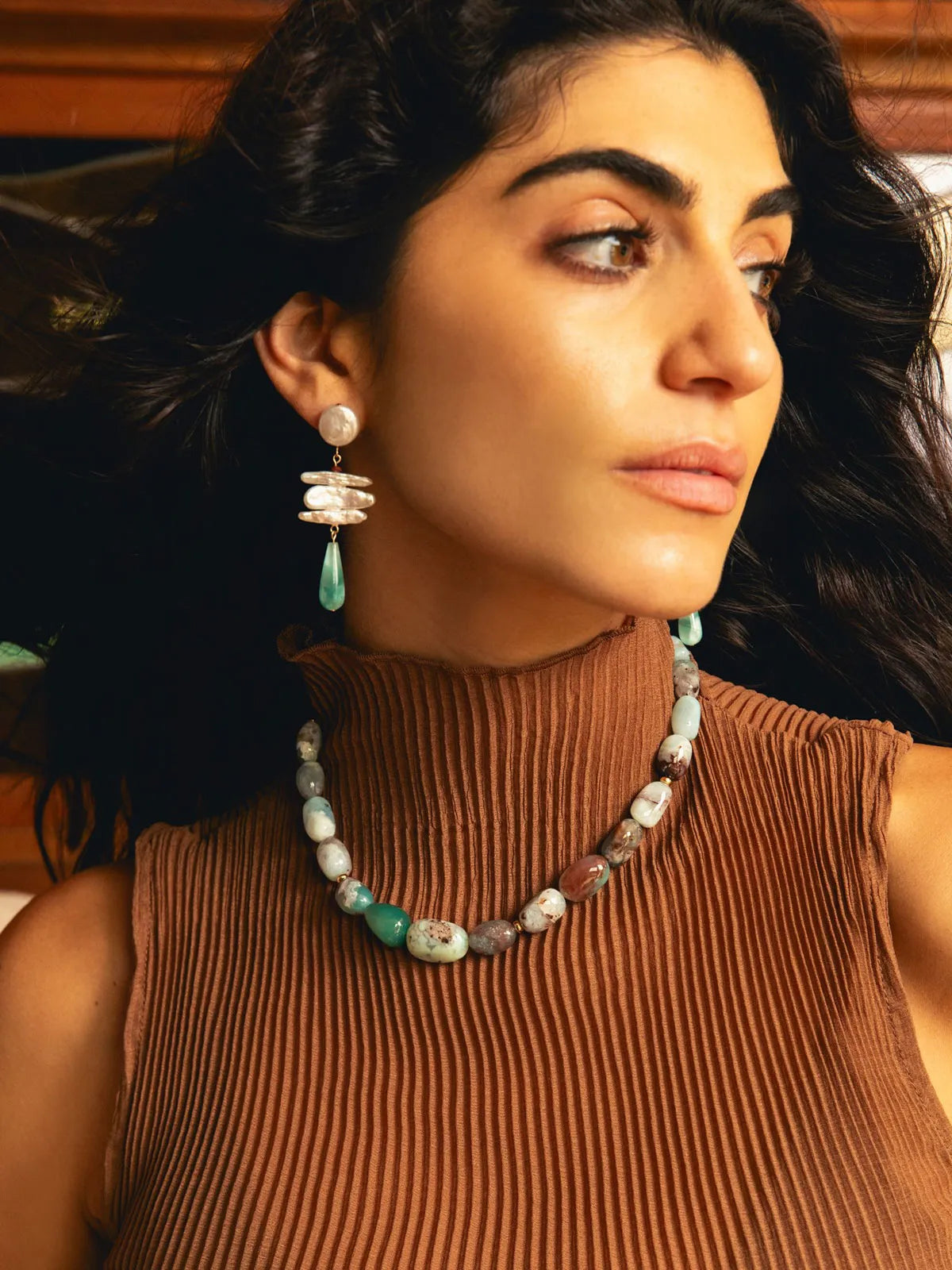 A woman wearing a brown ribbed turtleneck top and a chunky beaded necklace with turquoise and gray beads, along with matching drop earrings, shown in a warm indoor setting.